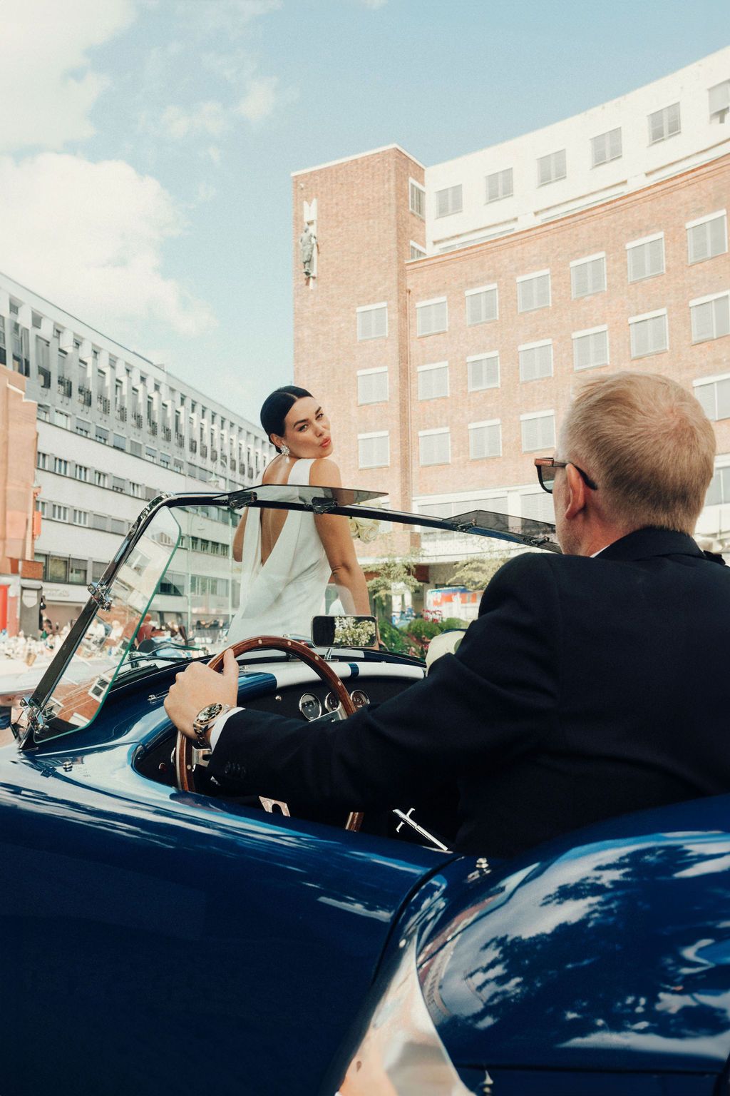 A vintage blue convertible car parked in front of a brick building during a wedding celebration.