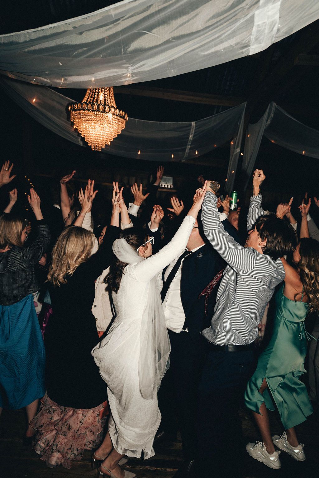 Wedding guests dancing with raised arms on a dark dance floor under draped fabric and crystal chandelier lighting.