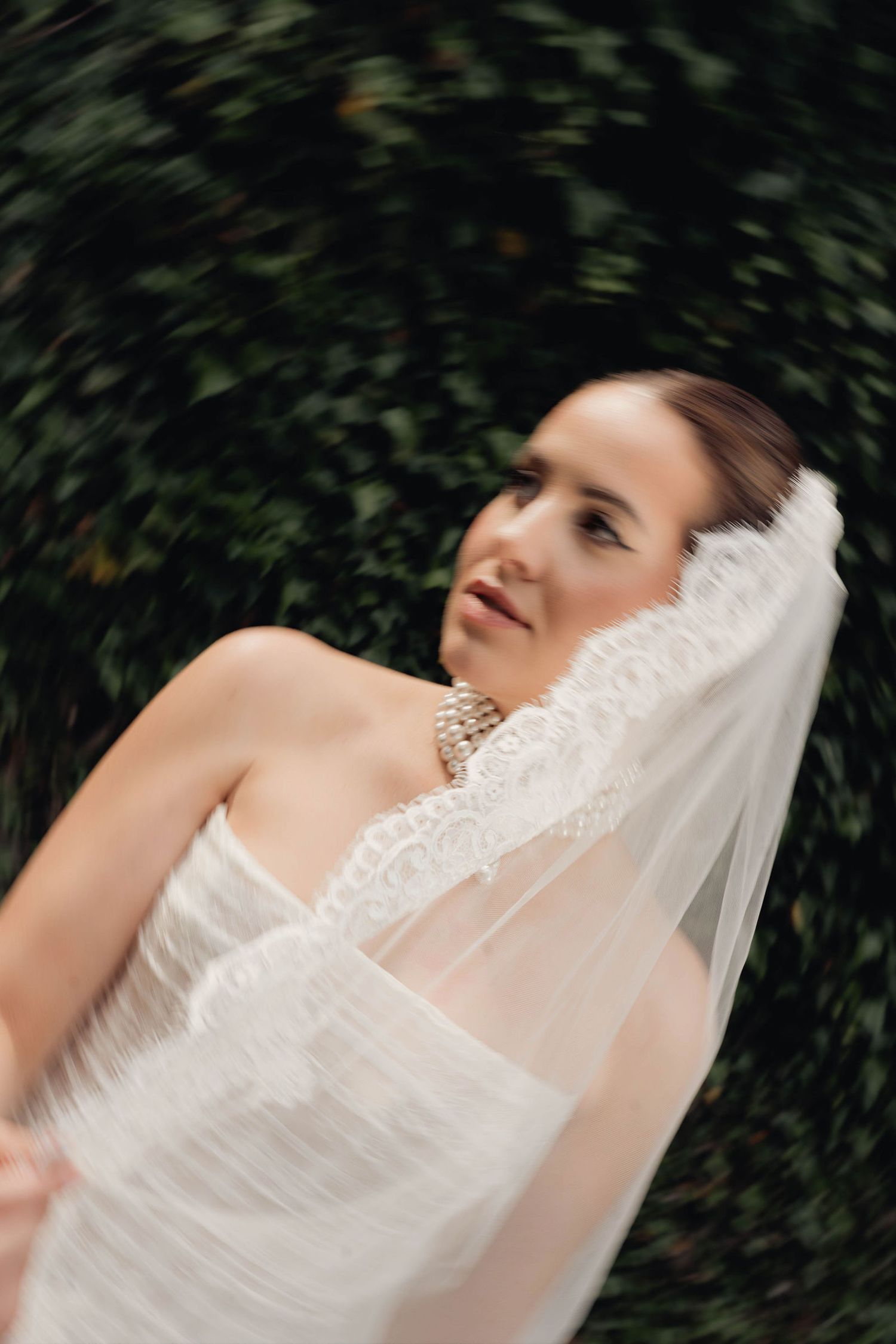 Bride in white strapless gown and lace-trimmed veil with pearl necklace, gazing upward against green foliage backdrop.