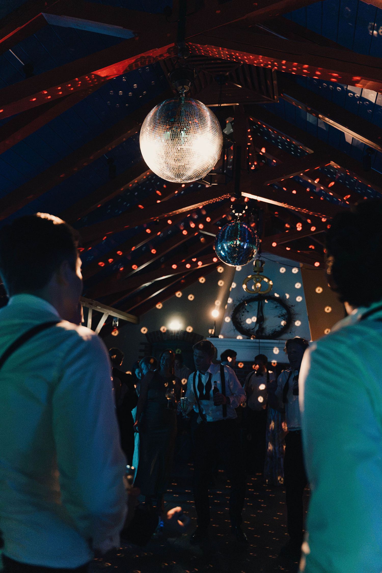 Disco ball glowing above a dimly lit dance floor with string lights and wooden ceiling beams at a party venue.