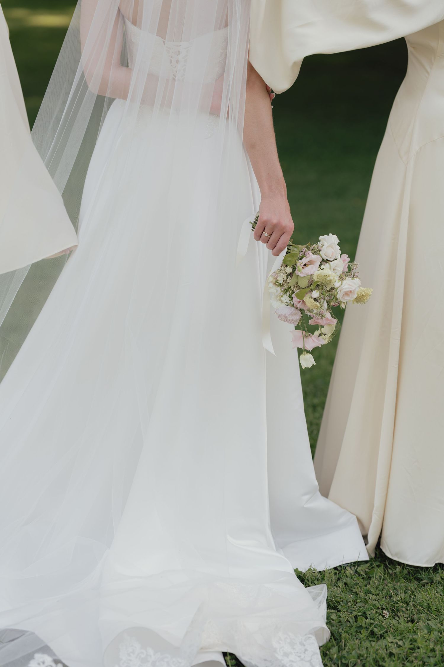 Bride in a flowing white gown and long tulle veil holds a pink and white bouquet on a green lawn beside a guest in a cream dress.