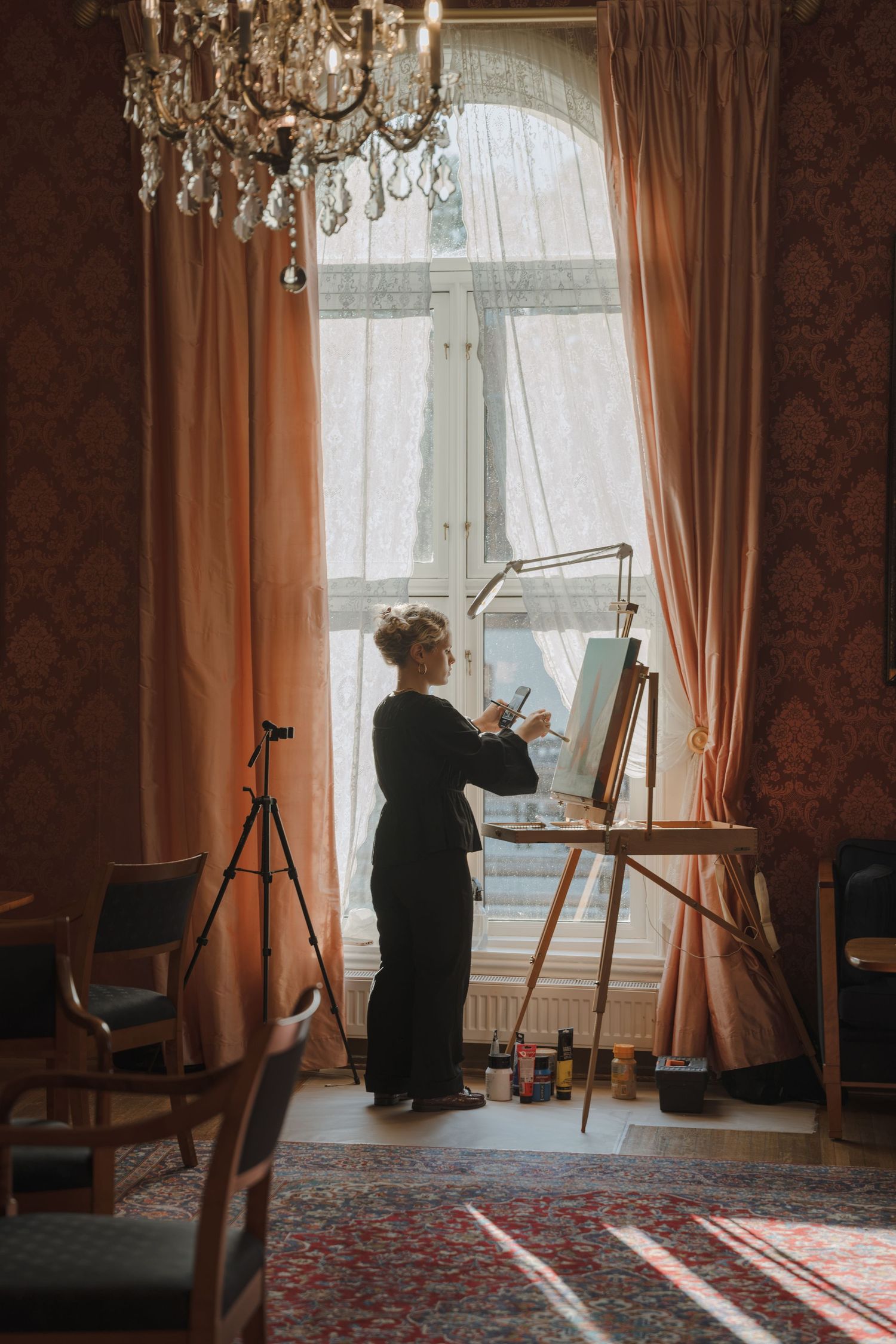 Woman in dark clothing paints at an easel by a tall arched window with peach curtains, chandelier, and patterned rug in an elegant room.