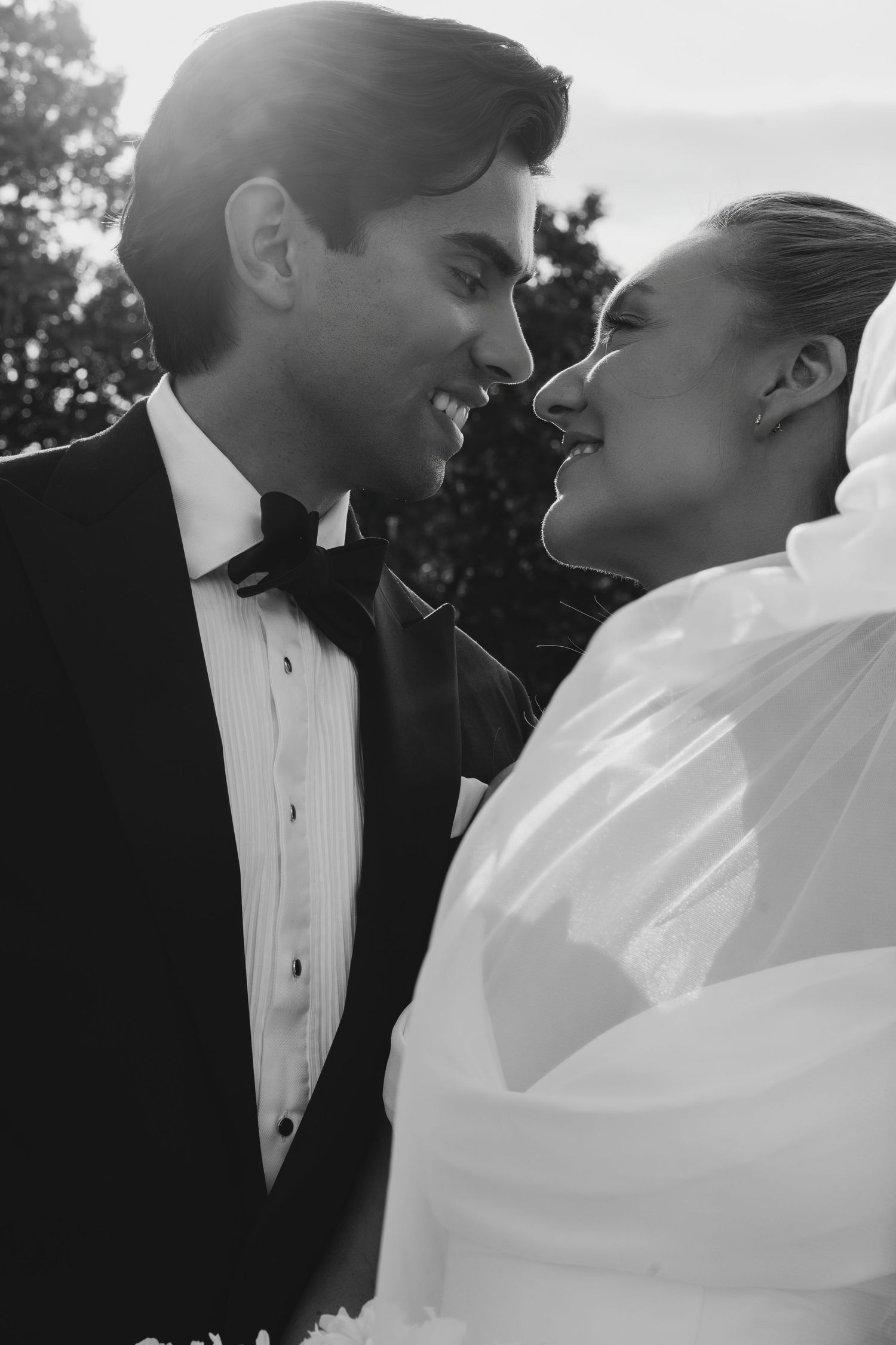 Black and white photo of a smiling bride in a flowing veil and a groom in a tuxedo and bow tie sharing a joyful moment outdoors.