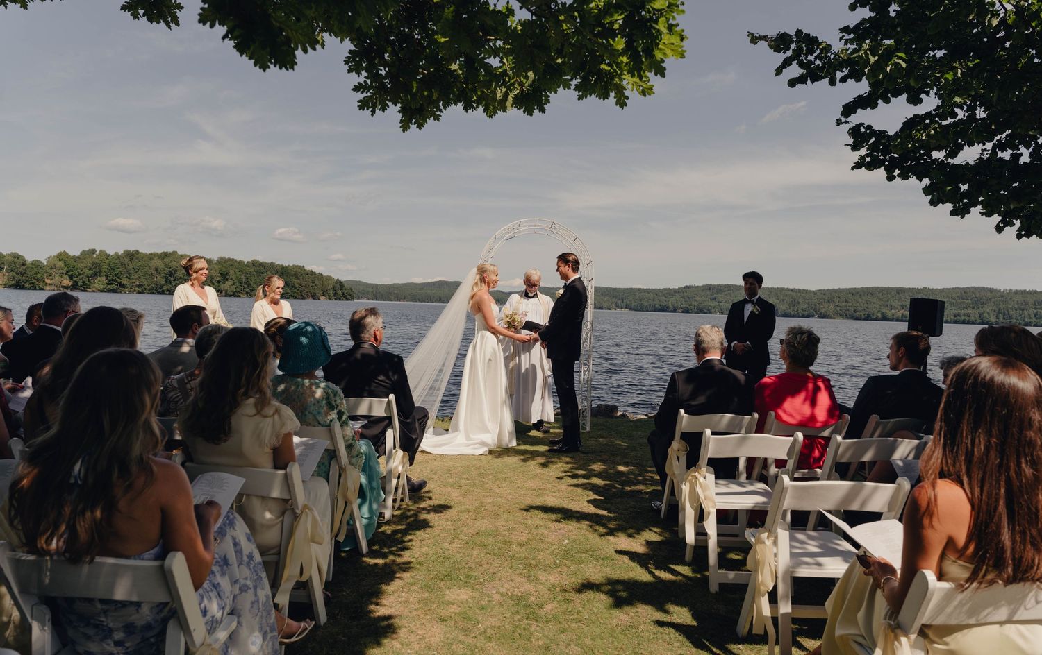Outdoor lakeside wedding ceremony with a bride and groom exchanging vows before seated guests on a grassy lawn surrounded by trees.