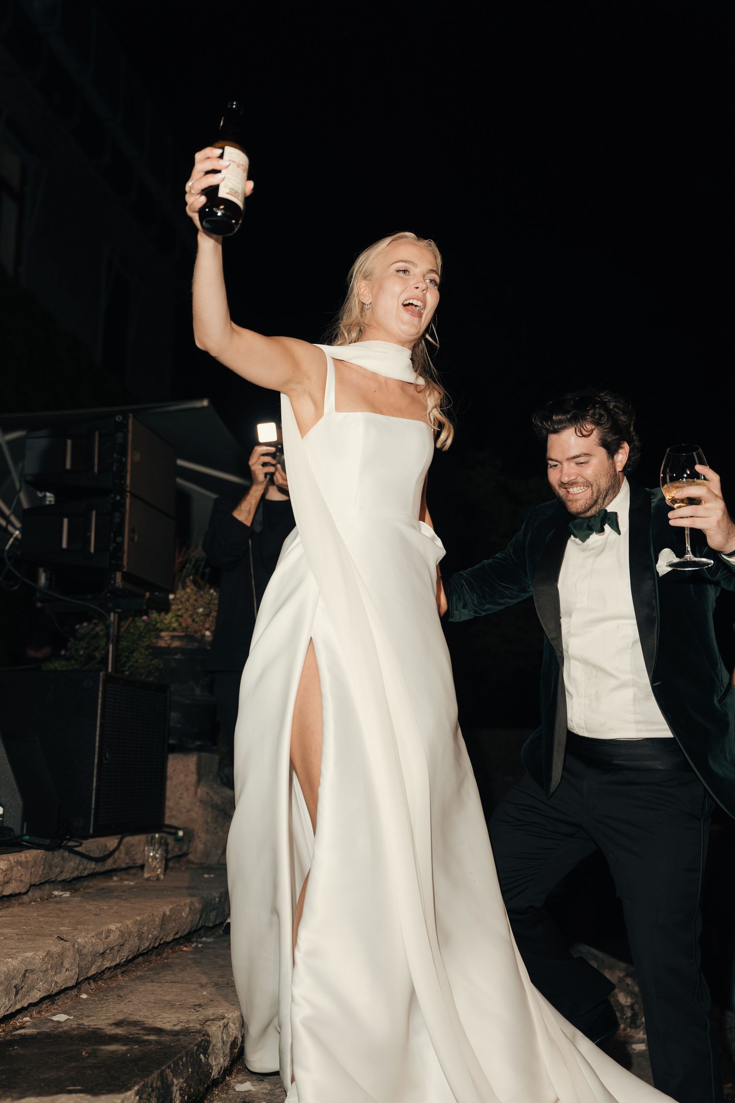 Bride in a white gown with a high slit raises a bottle in celebration beside a smiling groom holding a drink at night.