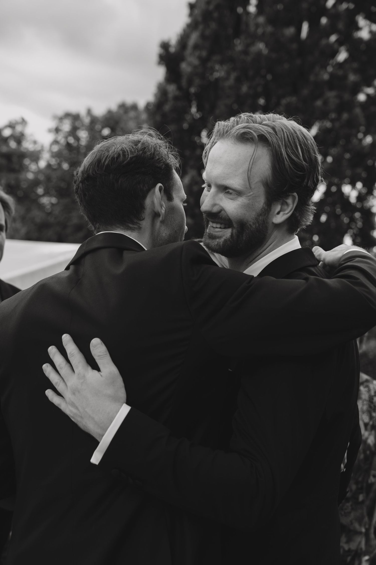 Black and white photo of two men in dark suits sharing a joyful hug outdoors at a wedding, trees in the background.