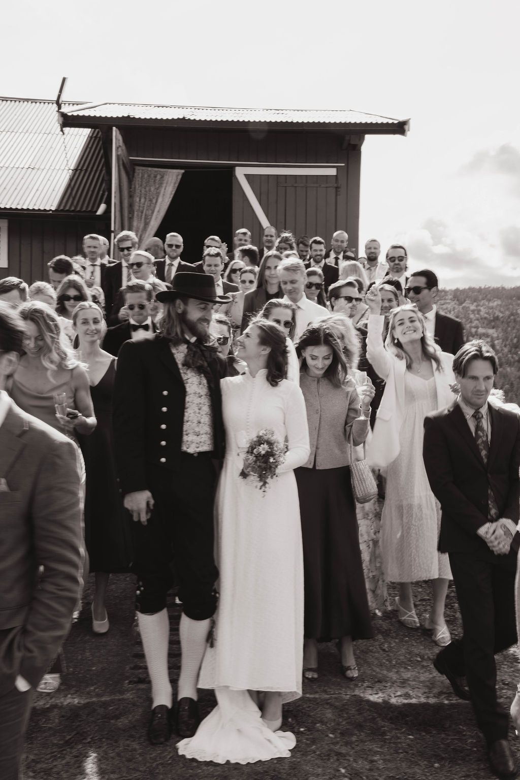 A black and white wedding party photo showing guests gathered outdoors near rustic wooden buildings.