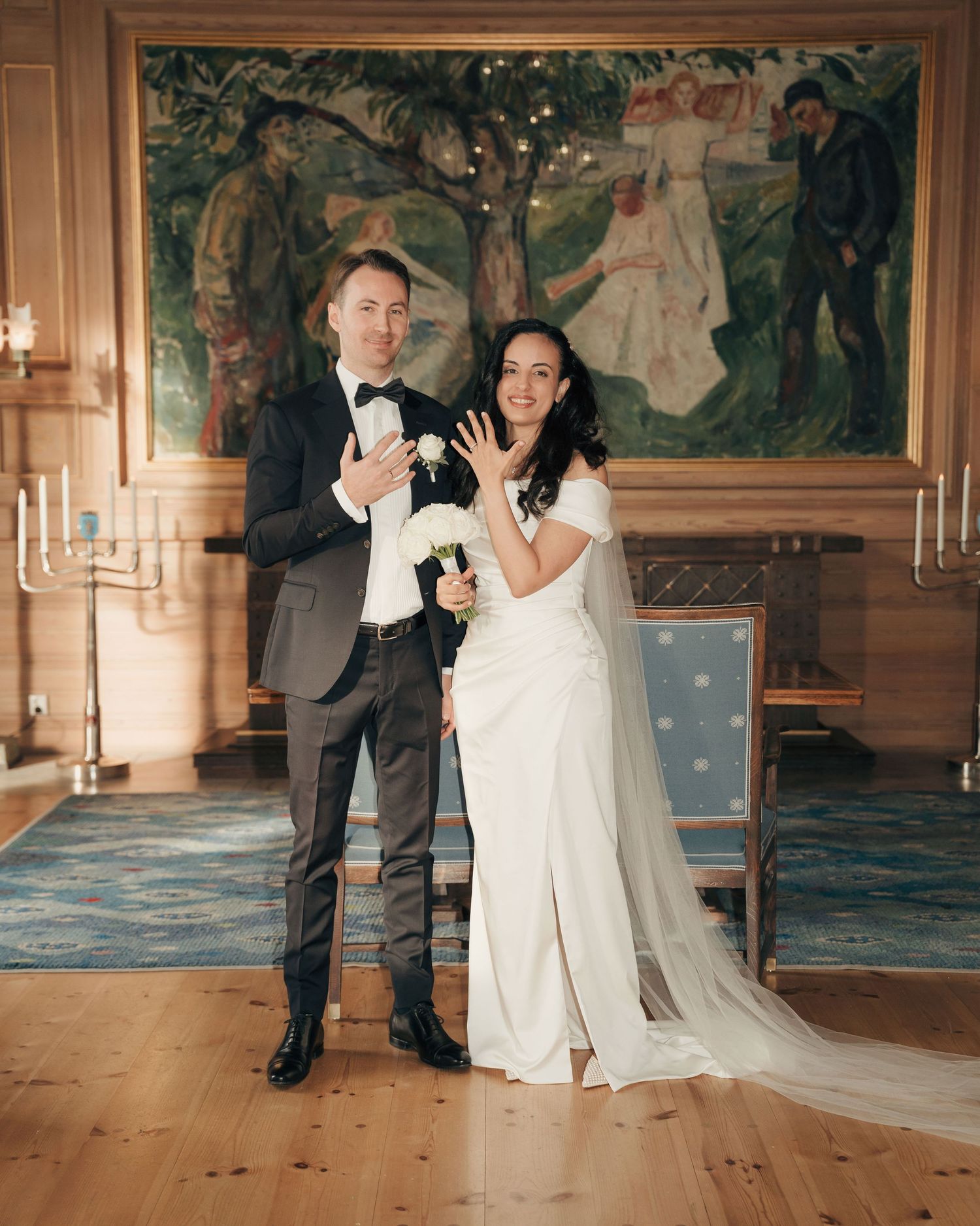 A couple in wedding attire shares a joyful moment in front of a decorative wall mural with candelabras in an elegant room.