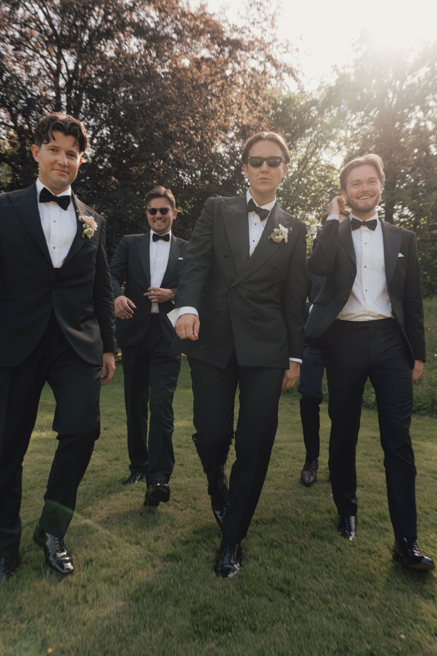 Four groomsmen in black tuxedos, bow ties and sunglasses stroll across a sunlit lawn with boutonnieres on their lapels.