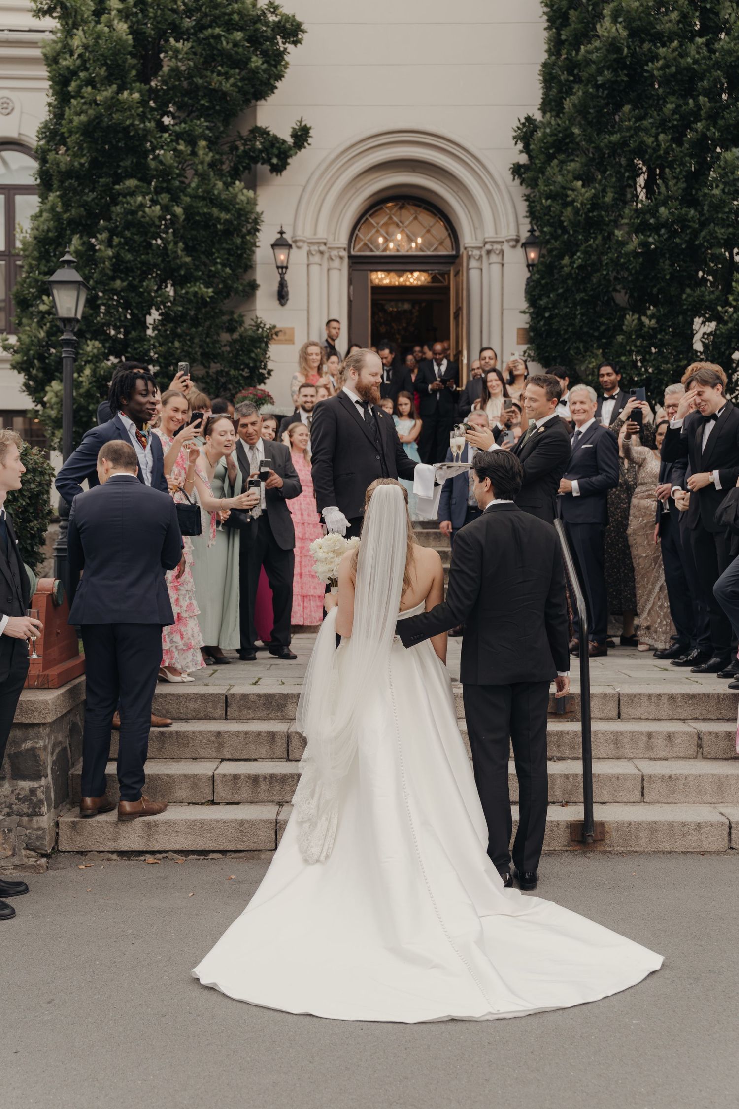 Bride in a long white gown and veil stands on stone steps facing smiling wedding guests in formal attire outside an arched church entrance.