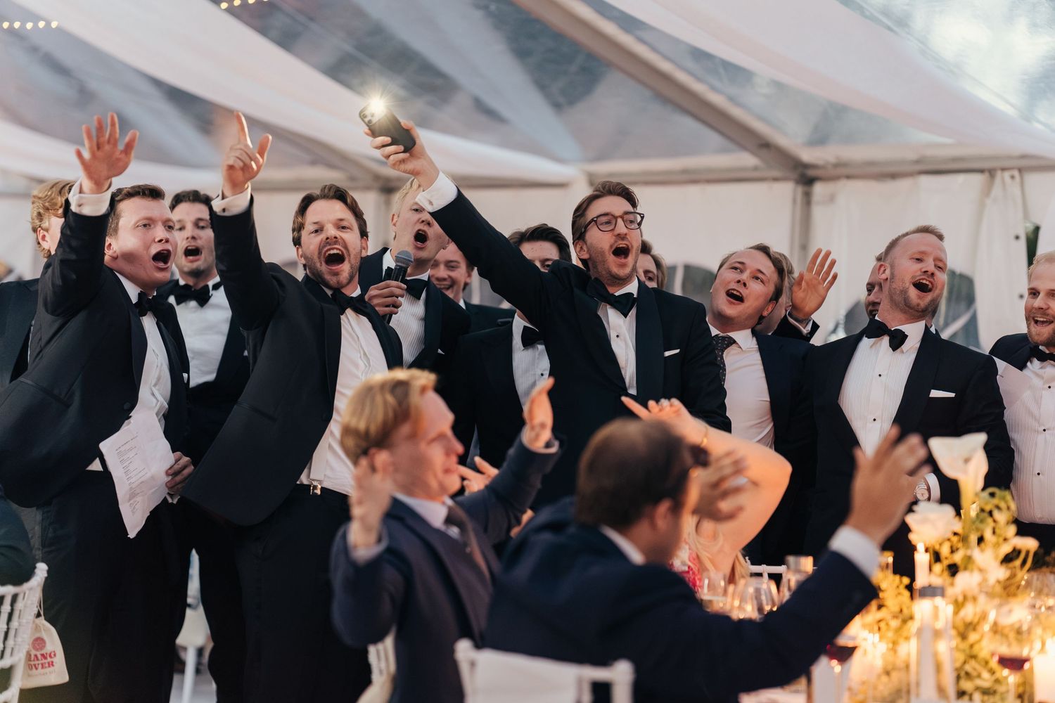 Groomsmen in black tuxedos cheer with raised arms and sing joyfully at a lively tented wedding reception.