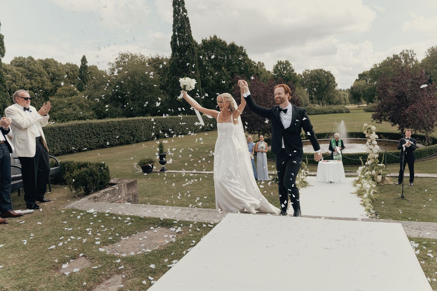 Bride and groom raise their arms in celebration while walking down a garden aisle as guests cheer and toss petals.