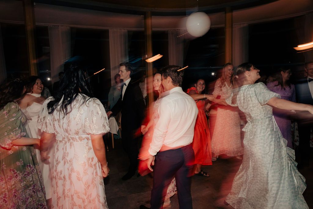 Blurred motion shot of people dancing at a dimly lit indoor wedding reception with red lighting accents.