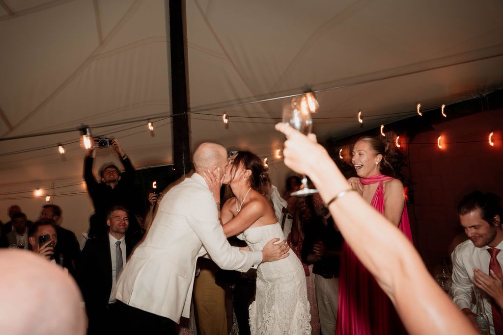 Wedding reception celebration under a tent with guests dancing and raising their hands during an energetic moment.