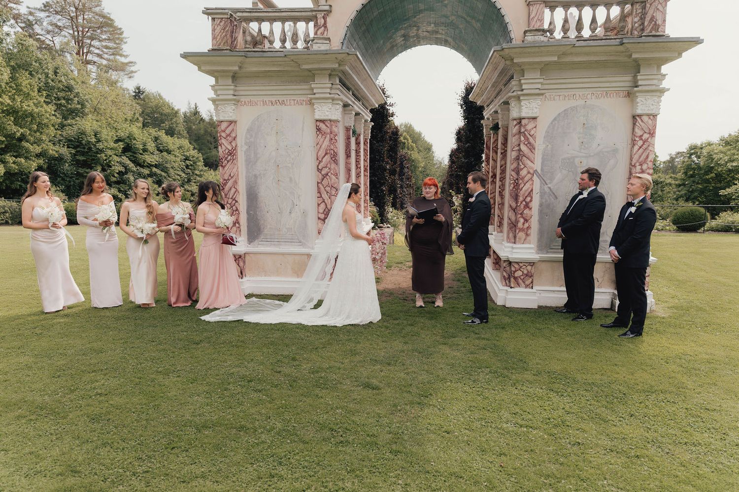 Outdoor wedding ceremony under ornate stone arch with bridesmaids in pink dresses and groomsmen on manicured lawn.