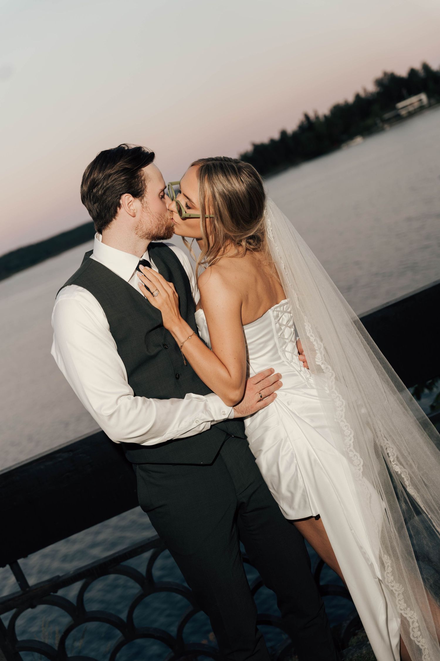 Bride and groom sharing romantic sunset kiss on waterfront dock wearing elegant wedding attire.
