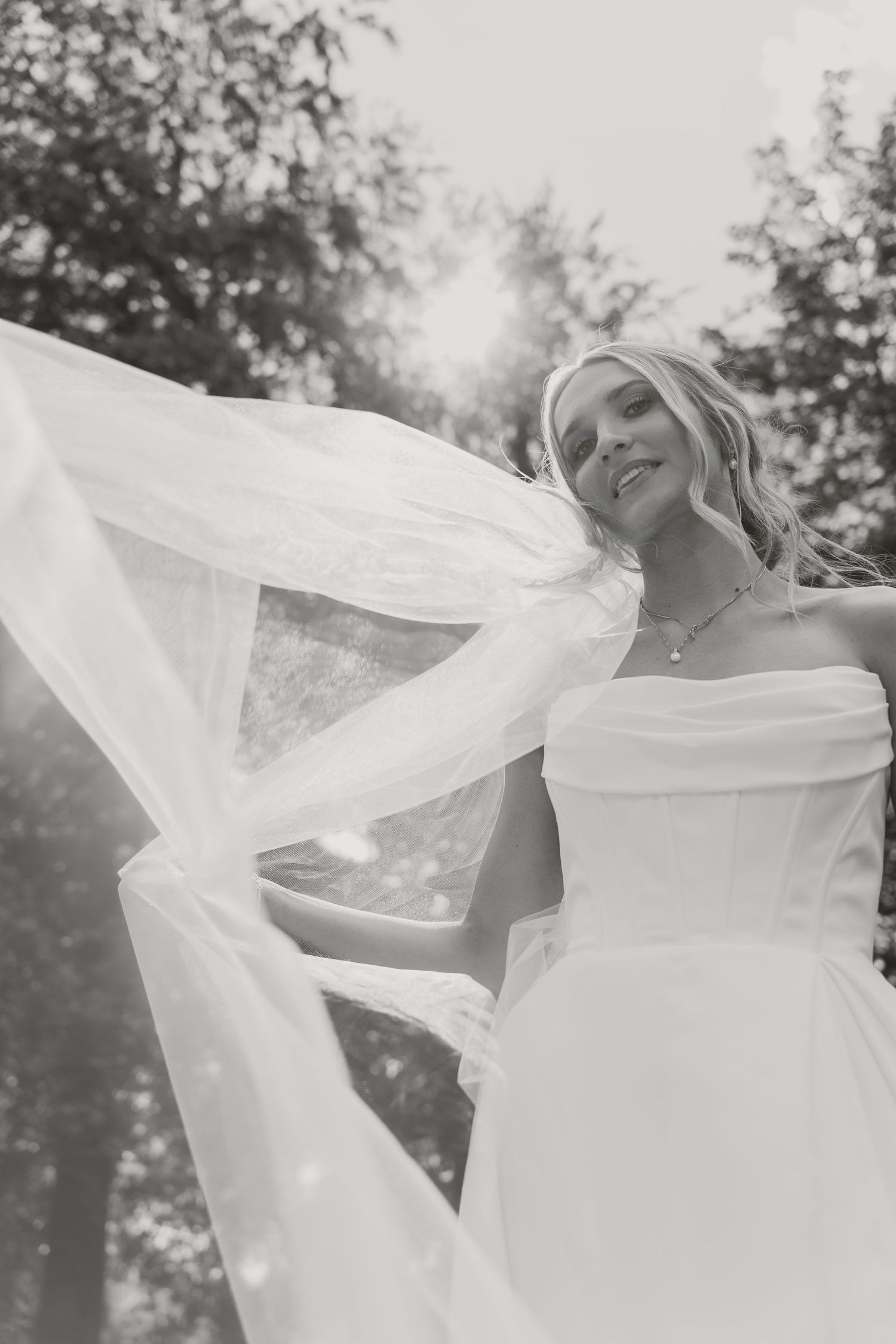 Black and white photo of smiling blonde bride in strapless gown with flowing veil billowing in the wind.