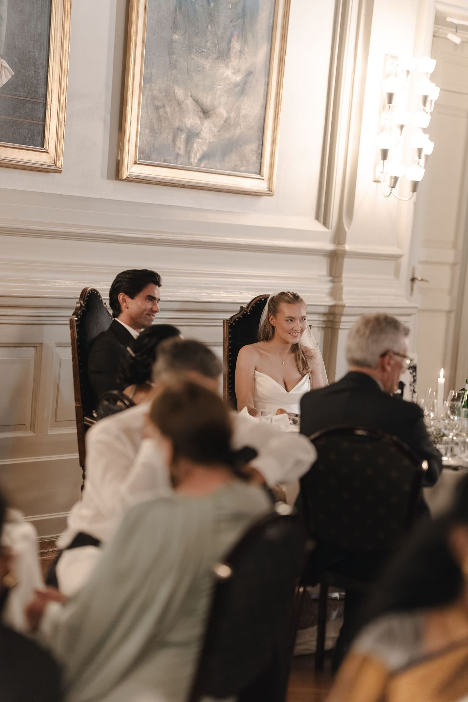 Smiling bride in a white dress and groom in a dark suit sit at an elegant wedding reception while guests watch in a classic ballroom.