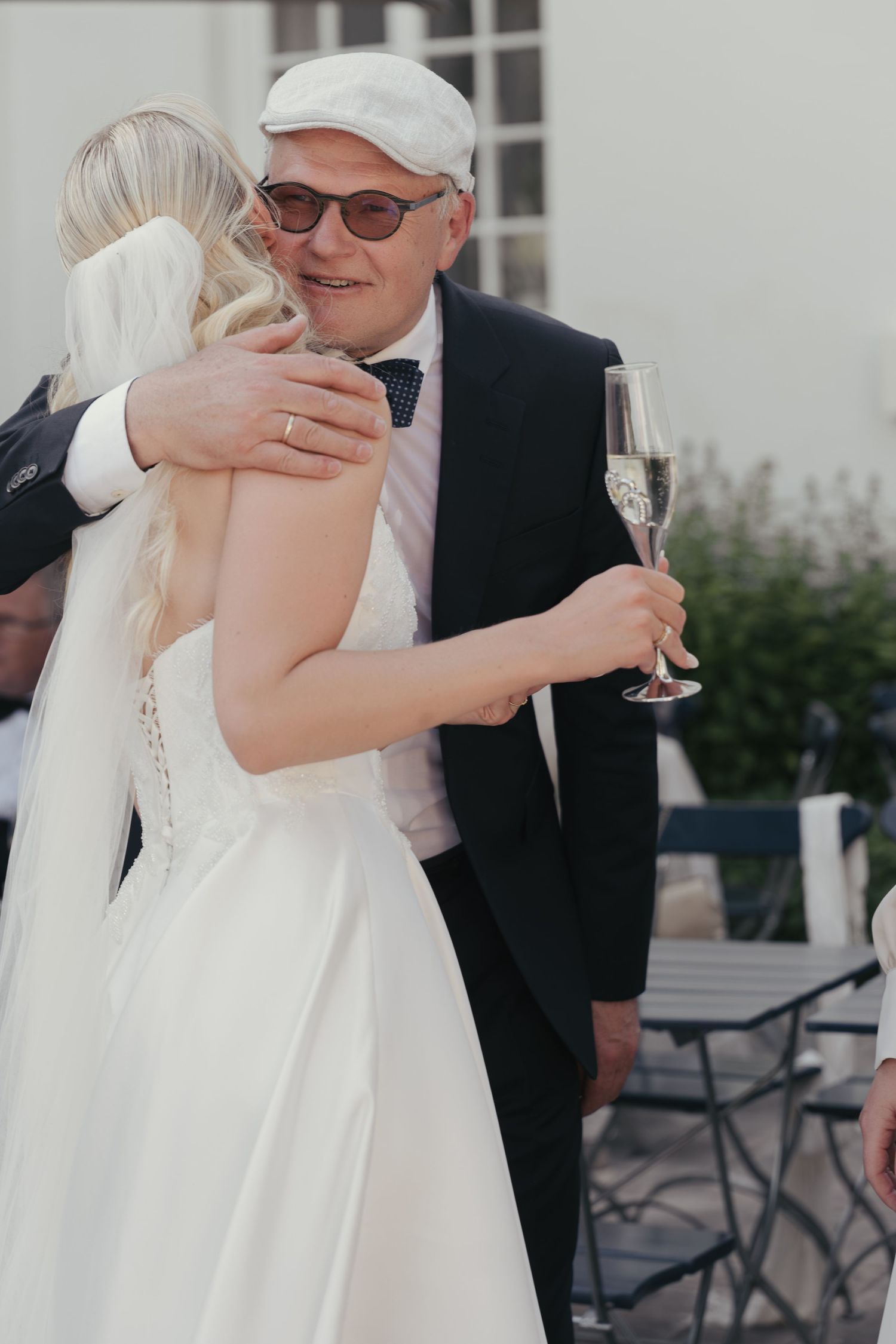 Bride in a white lace wedding gown and veil hugs an older man in a flat cap, sunglasses and bow tie as he holds a champagne glass.