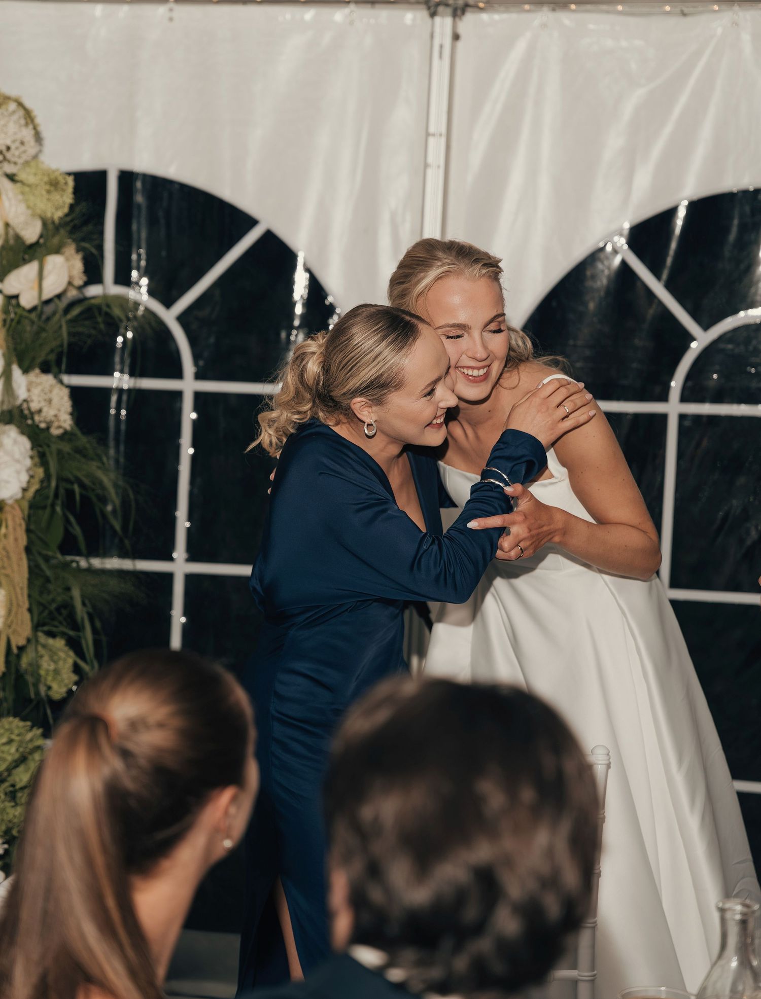 Bride in a white gown laughs and embraces a woman in a navy dress inside a decorated wedding tent with floral arrangements.