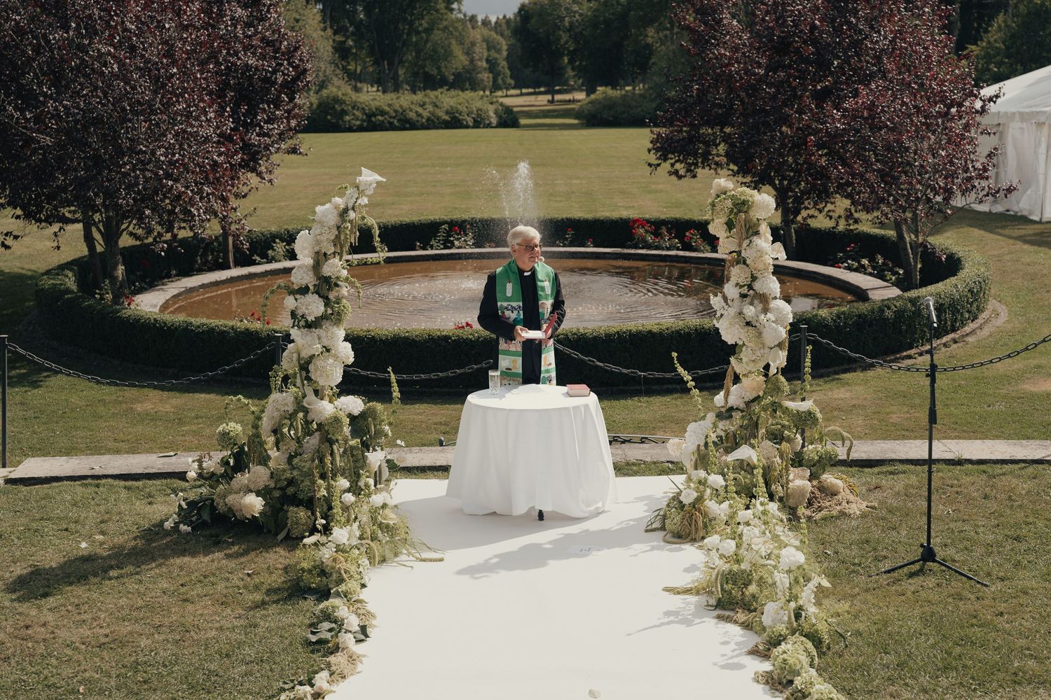 Officiant in a green stole stands at a white-draped table on an aisle lined with floral arrangements at an outdoor garden wedding ceremony.