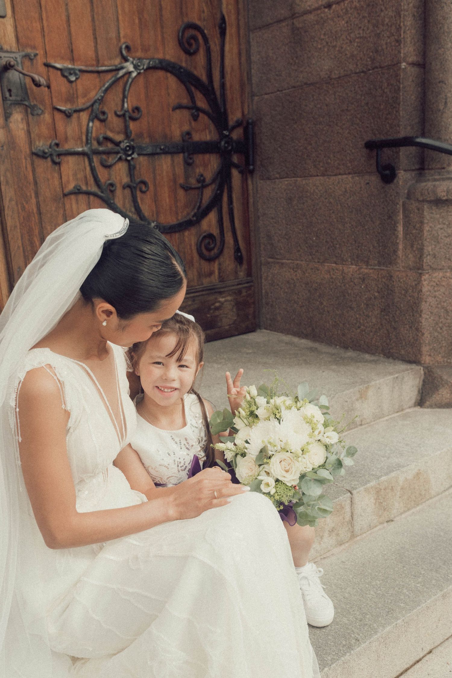 Bride and young flower girl in white dresses share a sweet moment on stone church steps.