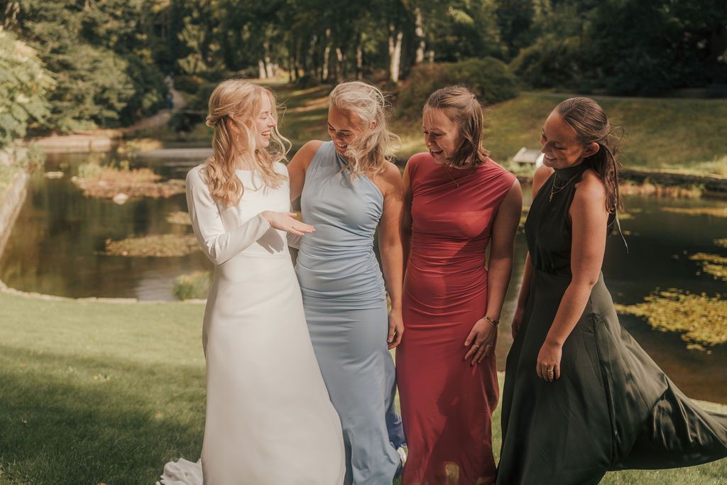 Four women in elegant formal dresses stand together outdoors near a pond on a sunny day.