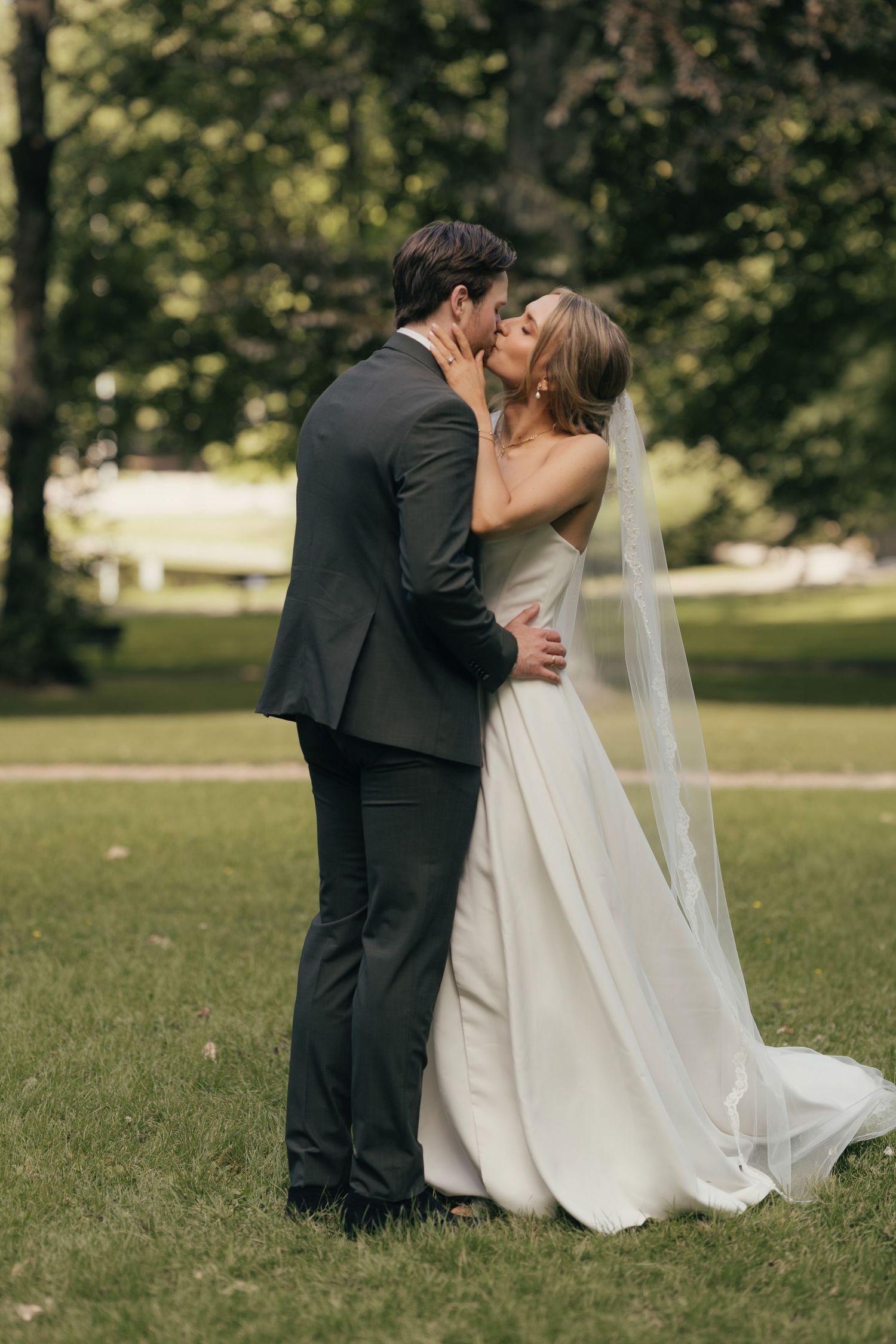 Bride and groom sharing a romantic kiss outdoors on green grass with trees in background.