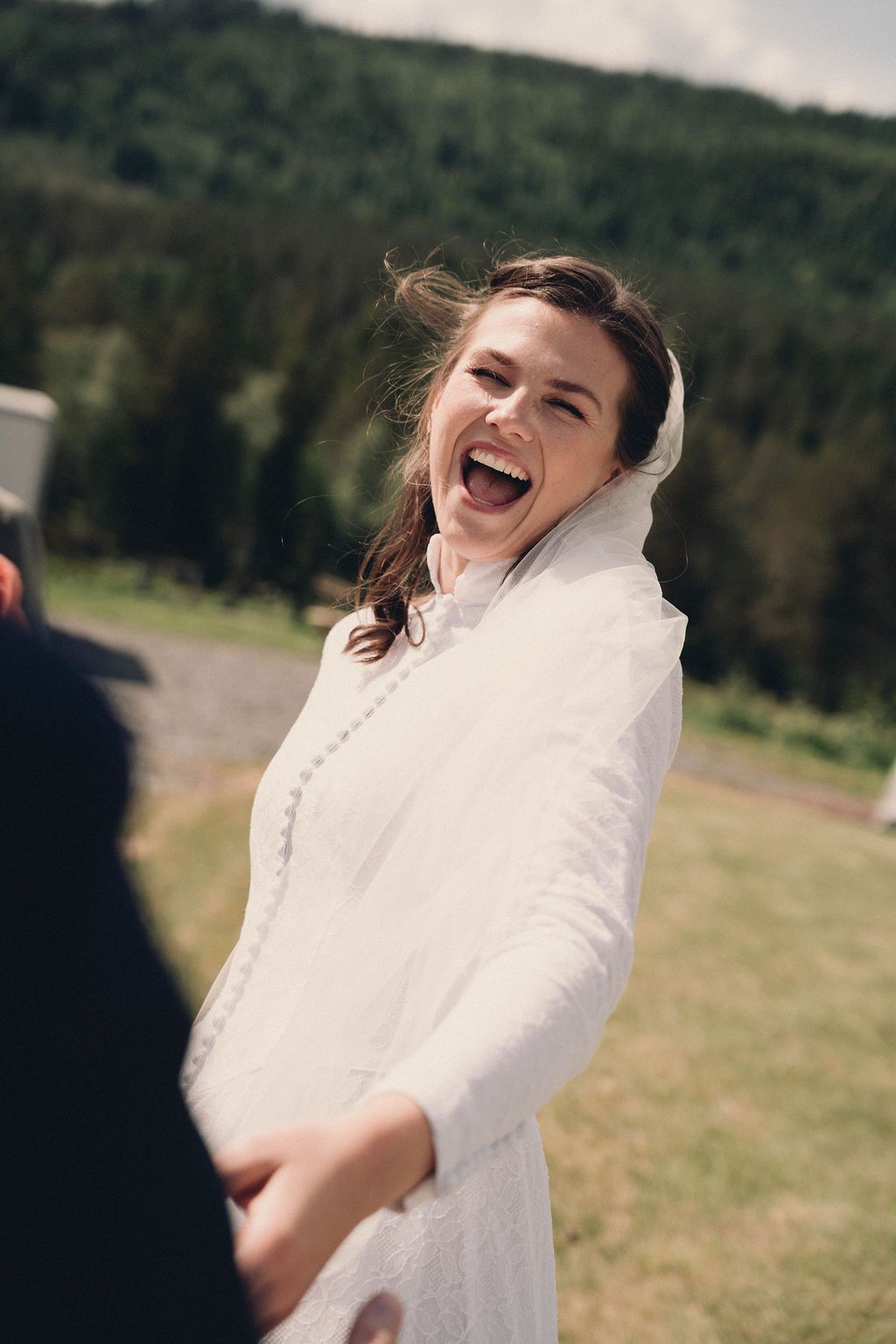A joyful bride in a white dress laughs candidly outdoors with mountains in the background.