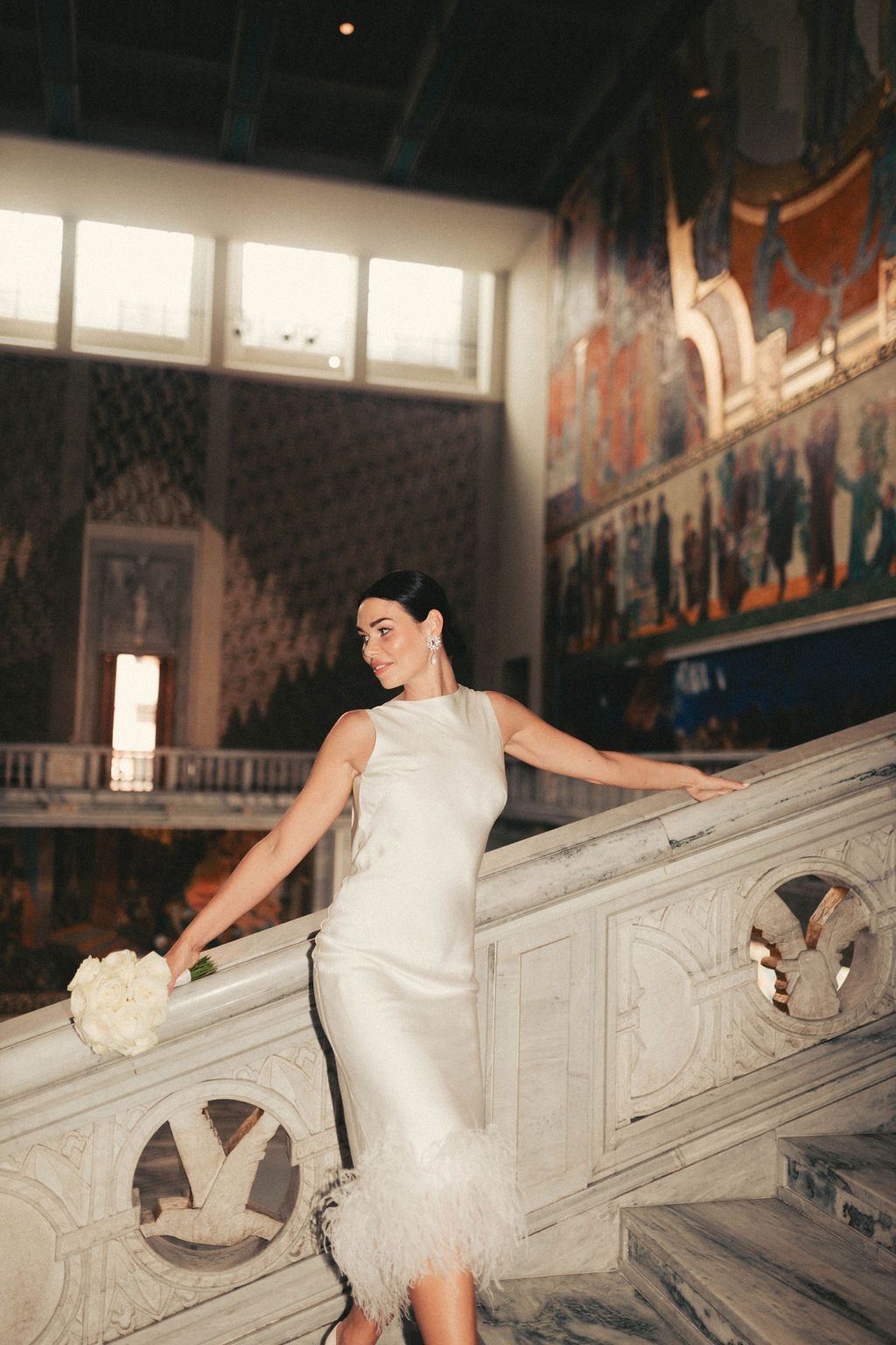 A bride in a white sleeveless dress poses gracefully on ornate marble steps in an elegant historic building.