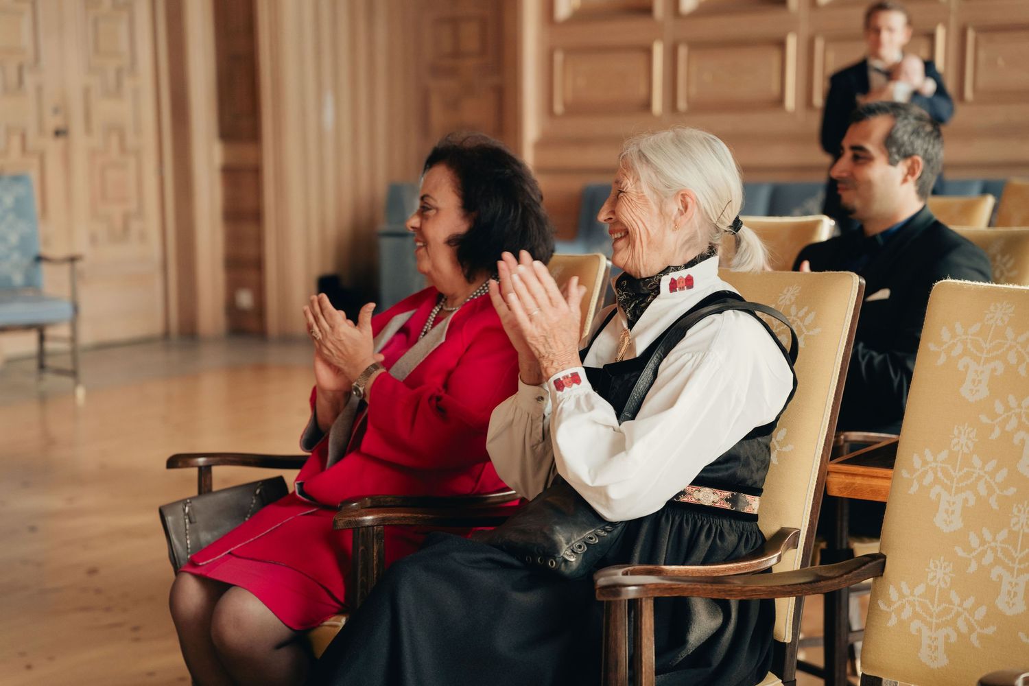People seated in a grand hall with wooden paneling applaud during a performance.