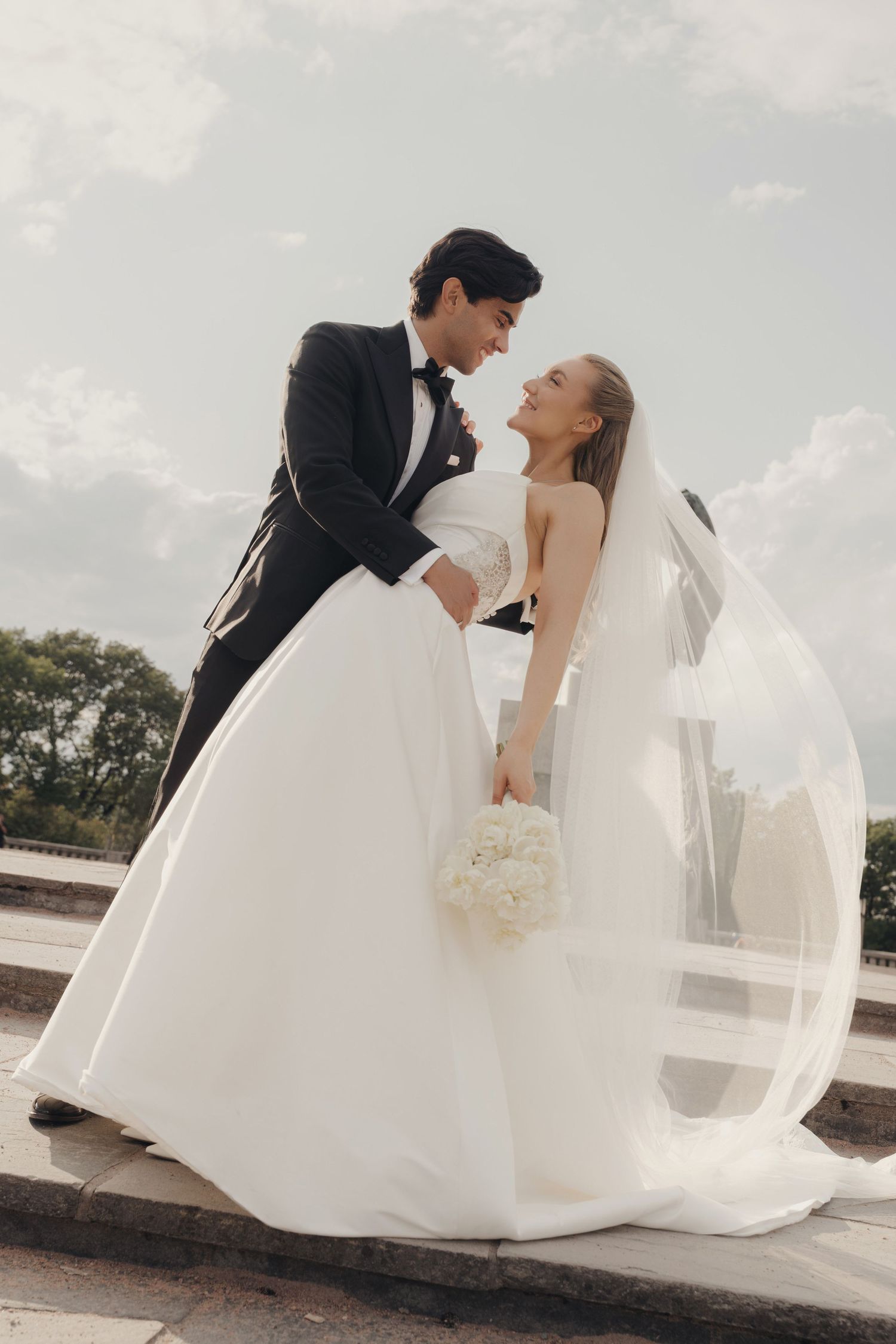 A bride in a strapless white gown and long veil holds a white bouquet as a groom in a black tuxedo dips her under a cloudy sky.