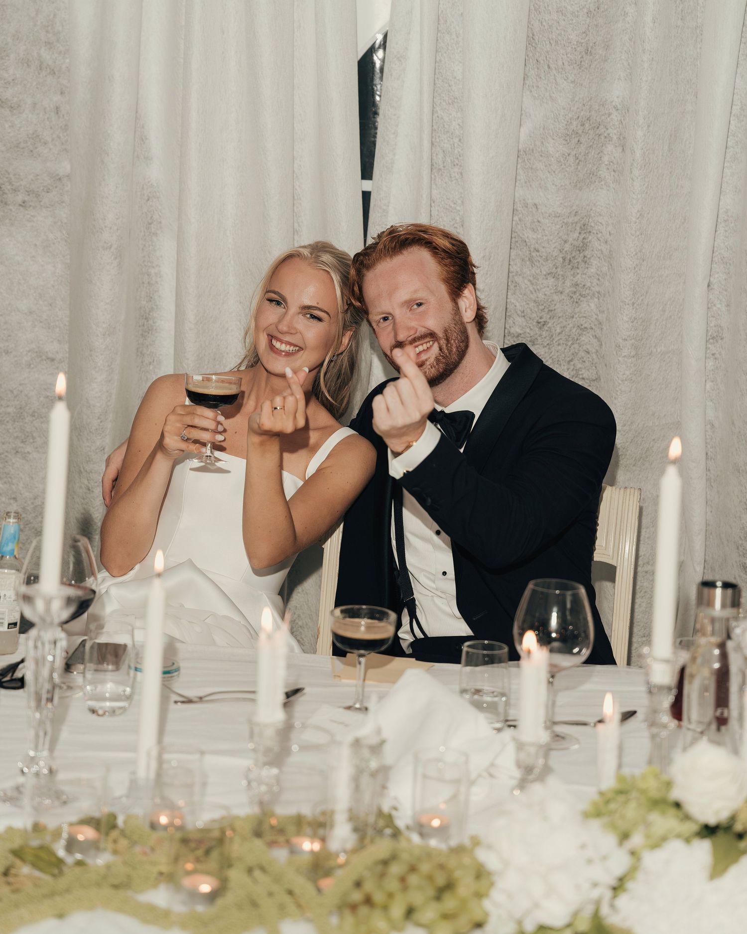 Smiling bride and groom raise glasses at a candlelit wedding reception table decorated with white flowers and greenery.