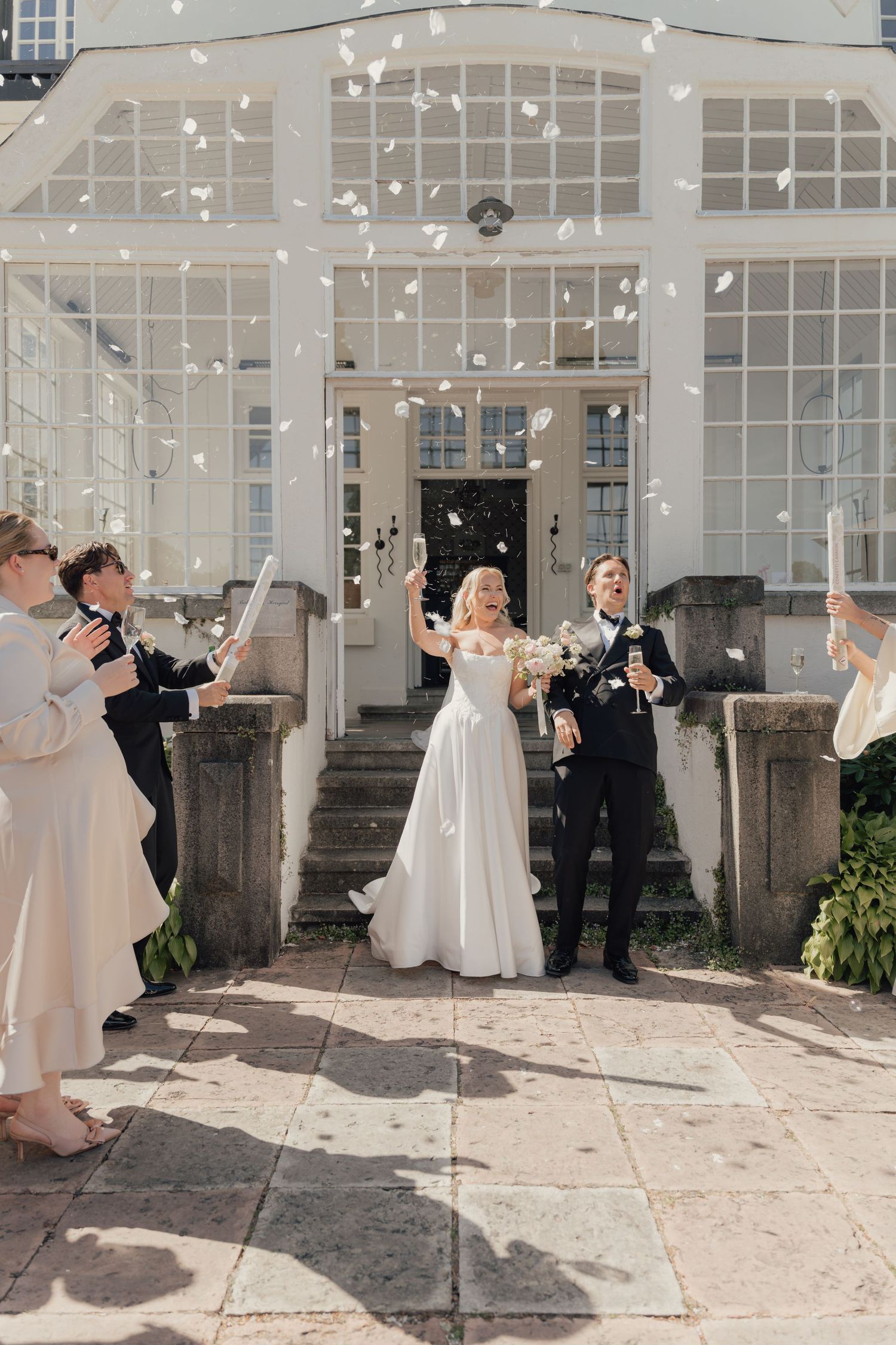 Newlyweds stand on stone steps in front of a glass conservatory as guests toss confetti and cheer after their wedding ceremony.