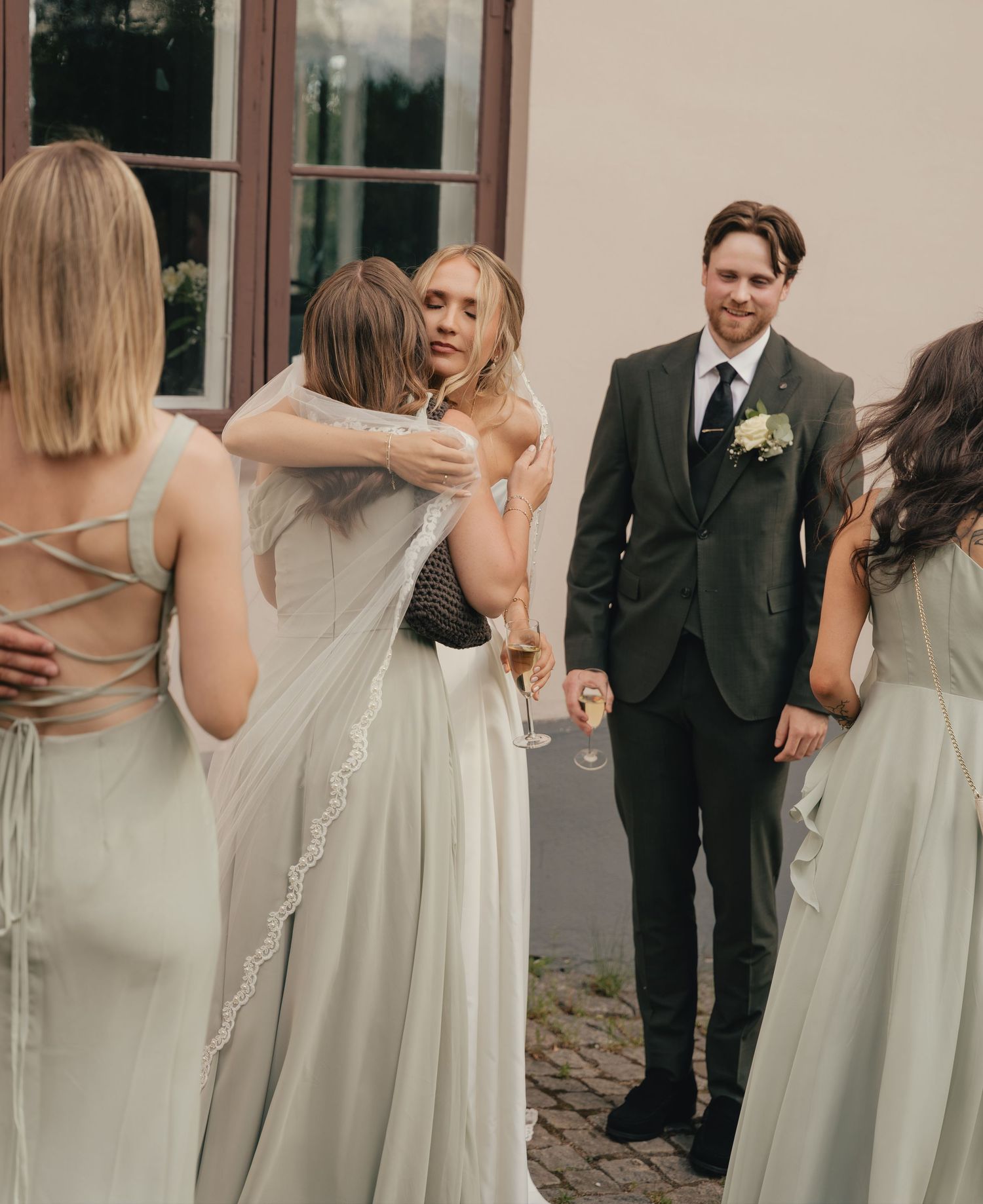 Bride hugging bridesmaid in sage green dress while groom watches during outdoor wedding reception.