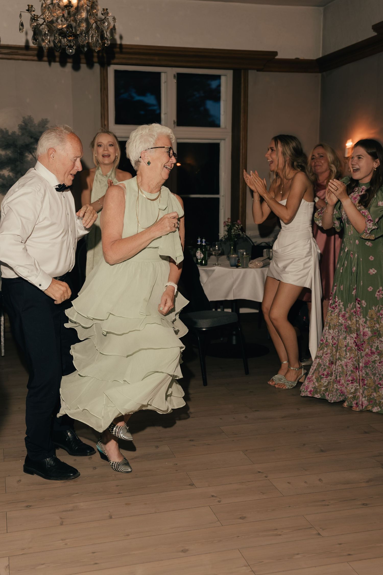 Elderly couple dancing energetically at wedding reception while guests cheer.