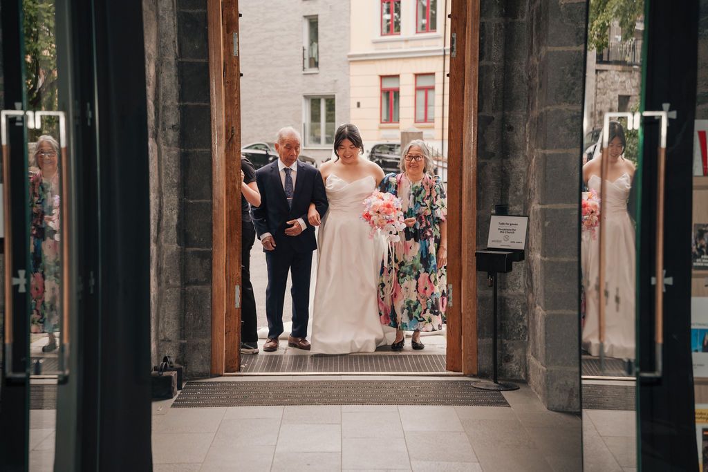 A wedding party walks through a stone archway in an old European city street during a celebration.