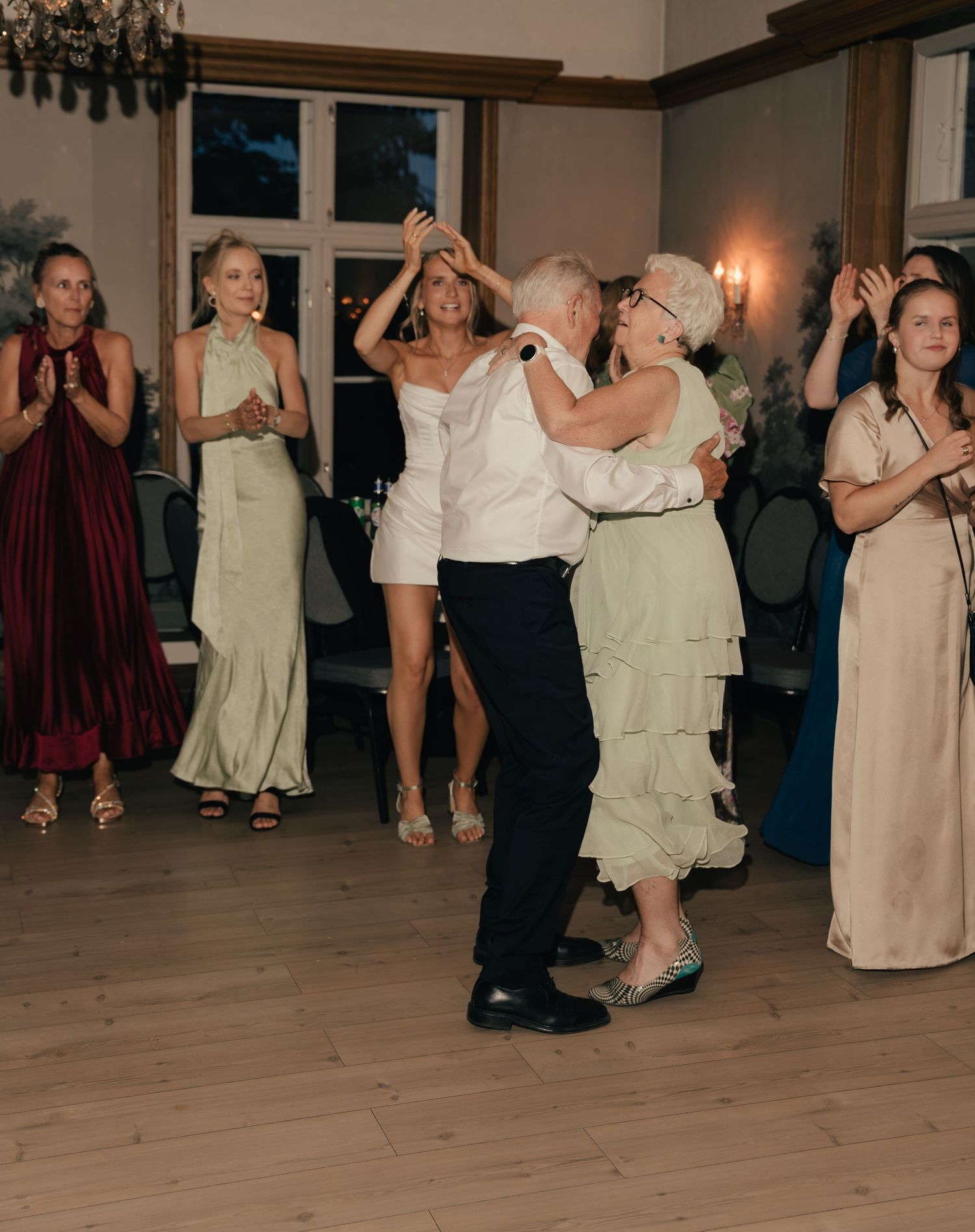 Elderly couple shares a dance on wedding reception floor surrounded by applauding guests.