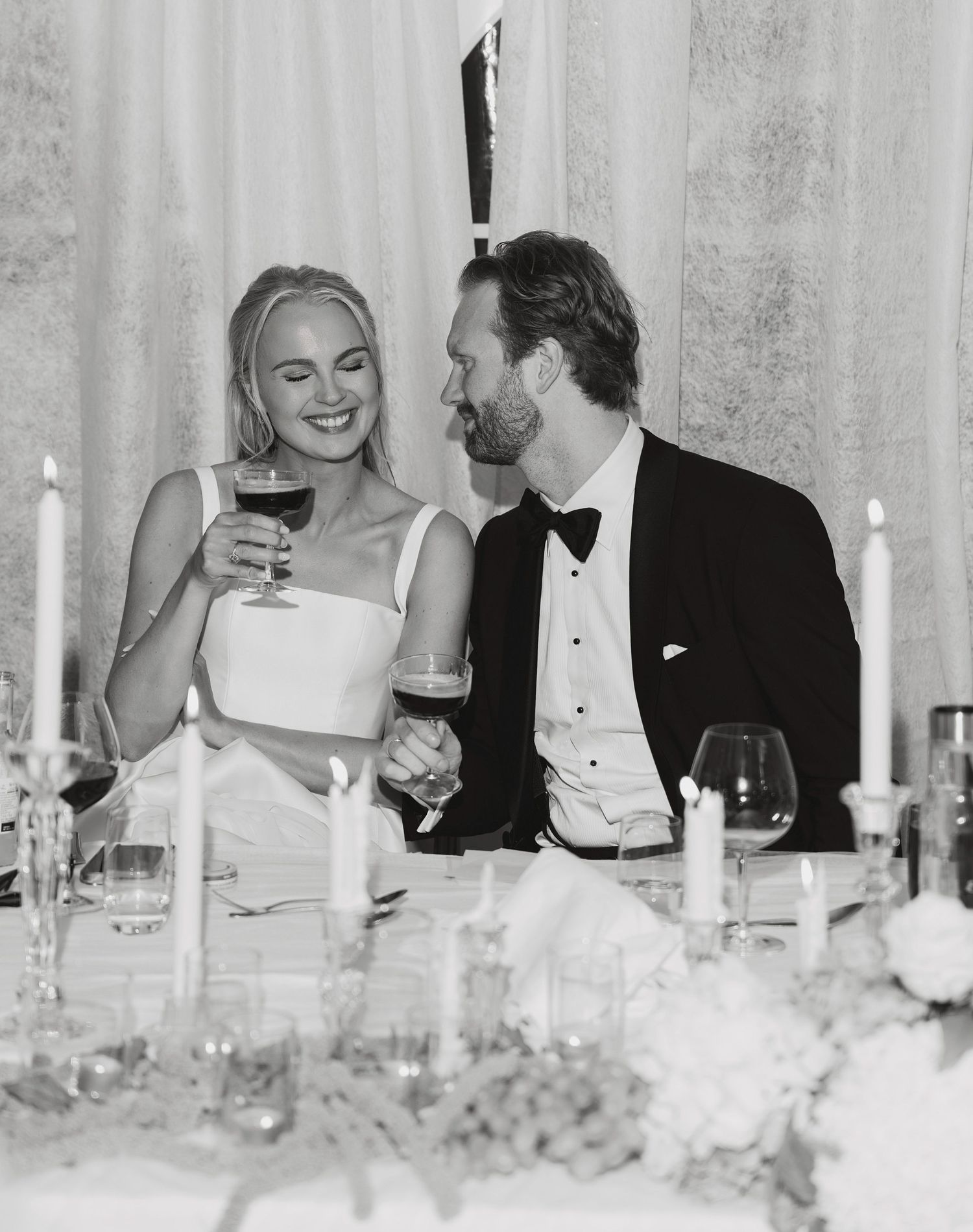 Smiling bride and groom in a black tuxedo raise wine glasses at a candlelit wedding reception table in black and white.