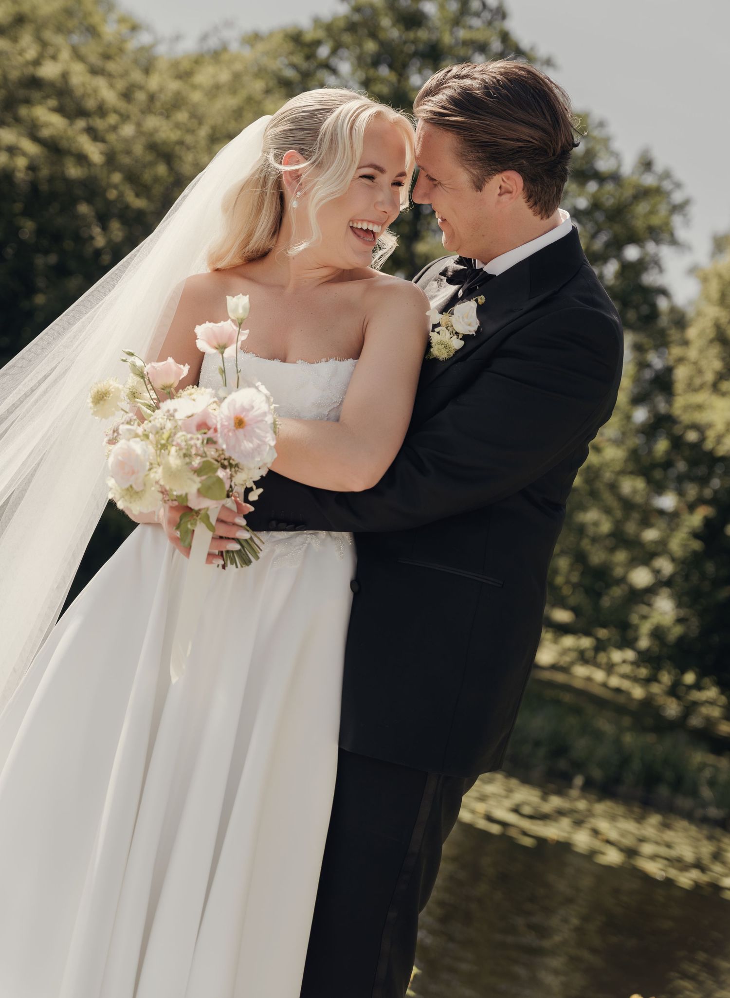 Laughing bride holding a pink and white bouquet embraces her groom in a black suit beside a calm lake.