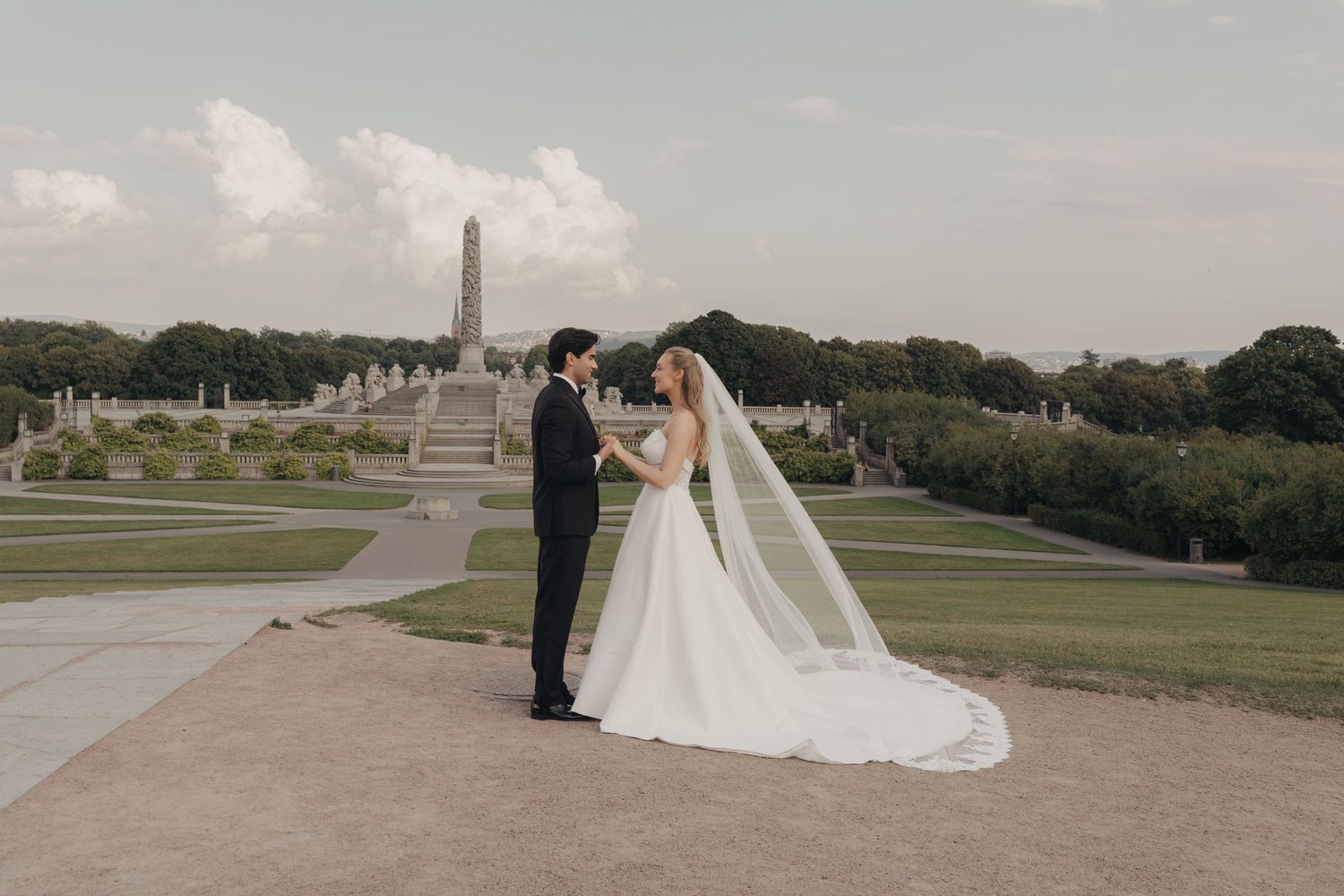 Bride and groom embrace in a park with a long lace veil, a stone monument and sculpted gardens behind them.