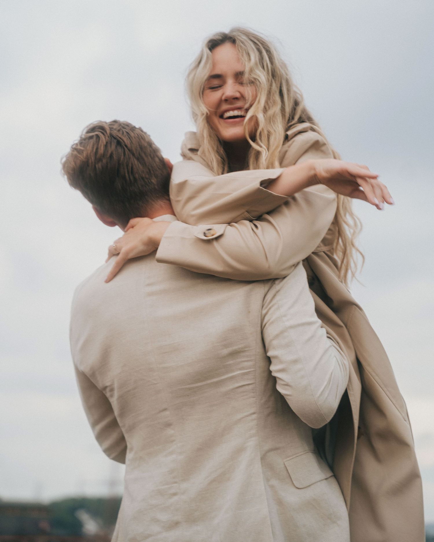 A joyful couple embraces outdoors wearing matching beige coats against an overcast sky.
