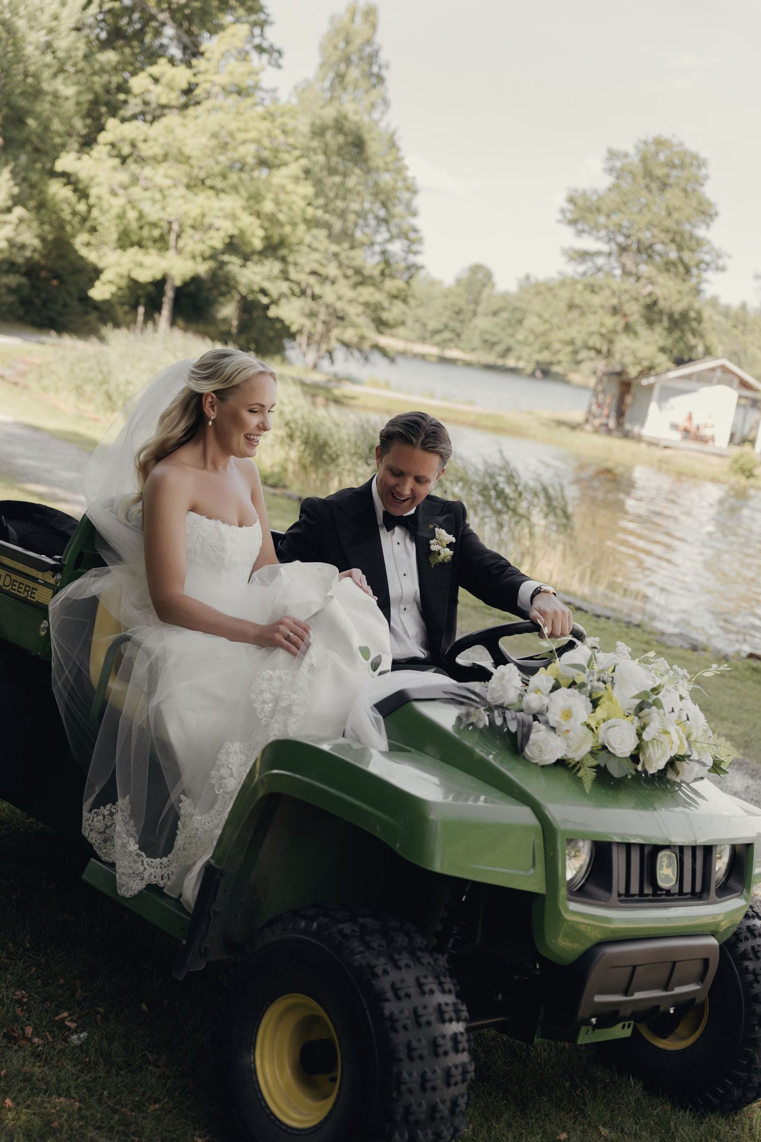 Smiling bride in a lace wedding gown and groom in a tuxedo ride a green utility vehicle with a white floral bouquet by a lakeside.