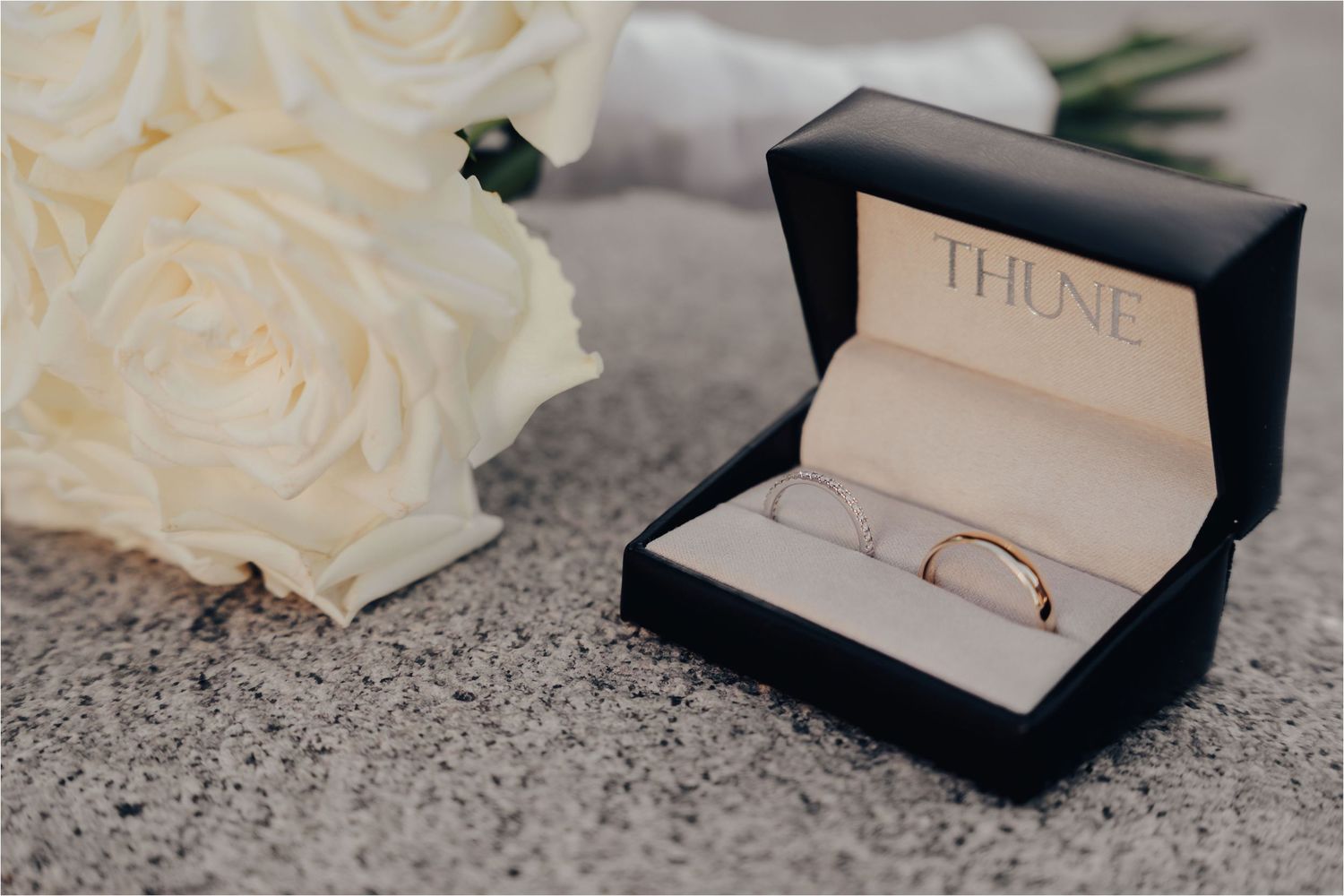A gold wedding ring sits in a black Thune jewelry box next to white roses on a textured gray surface.