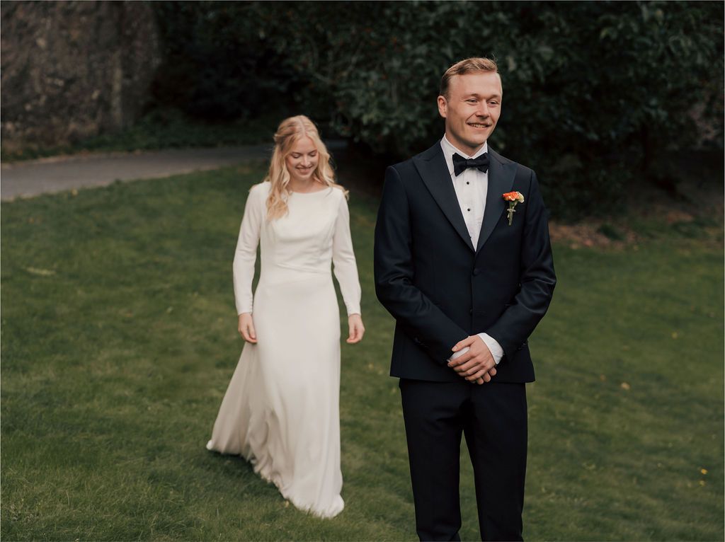 A bride and groom stand on a green lawn during their outdoor wedding, with the bride wearing a long-sleeved white gown.