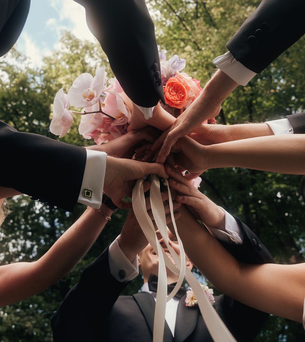 Multiple hands come together in a circle wearing corsages and holding pink flowers in an outdoor garden setting.