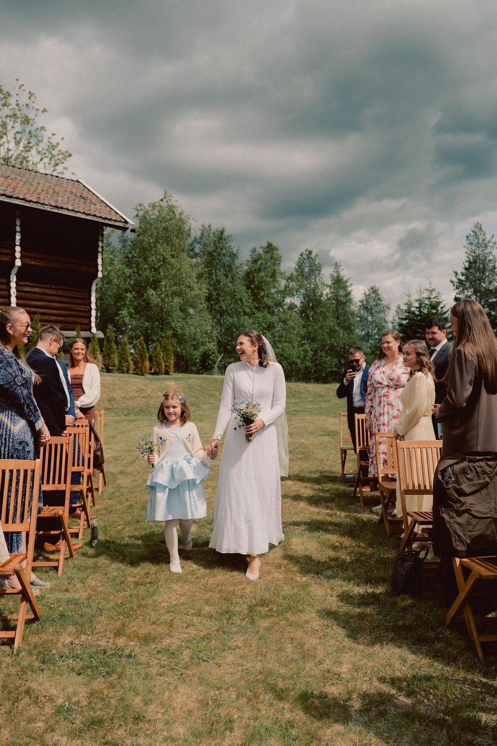 Guests gather on wooden chairs at an outdoor rustic wedding ceremony with a log cabin and trees in the background.