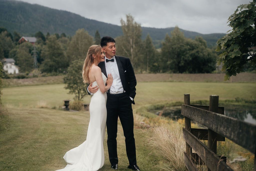 A romantic wedding couple embraces on a scenic countryside hillside with a rustic wooden fence in the background.