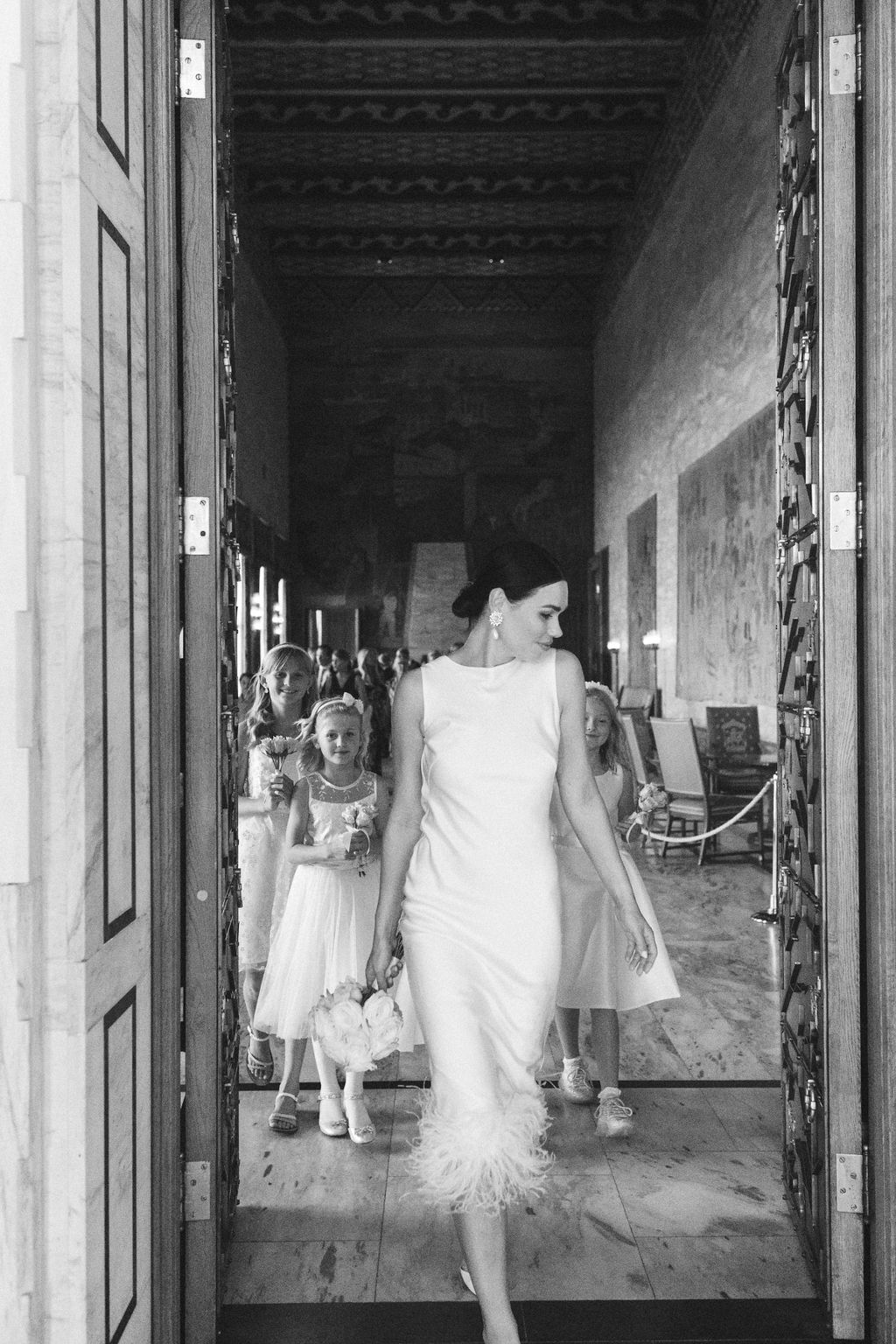 A black and white photograph showing a bride in a sleeveless dress walking through an elegant hallway.