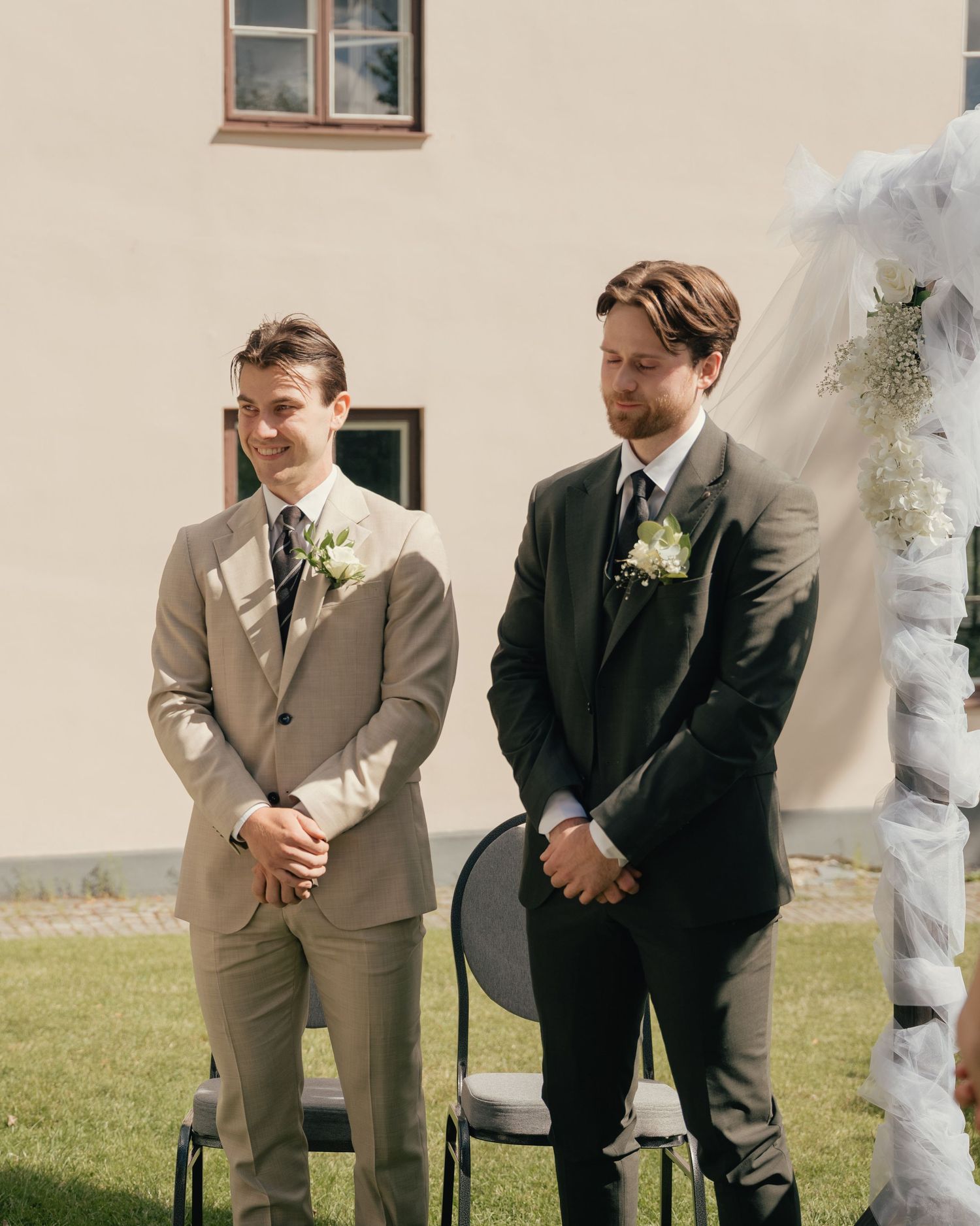 Two grooms stand at outdoor wedding altar wearing suits with boutonnieres, smiling joyfully on sunny day.